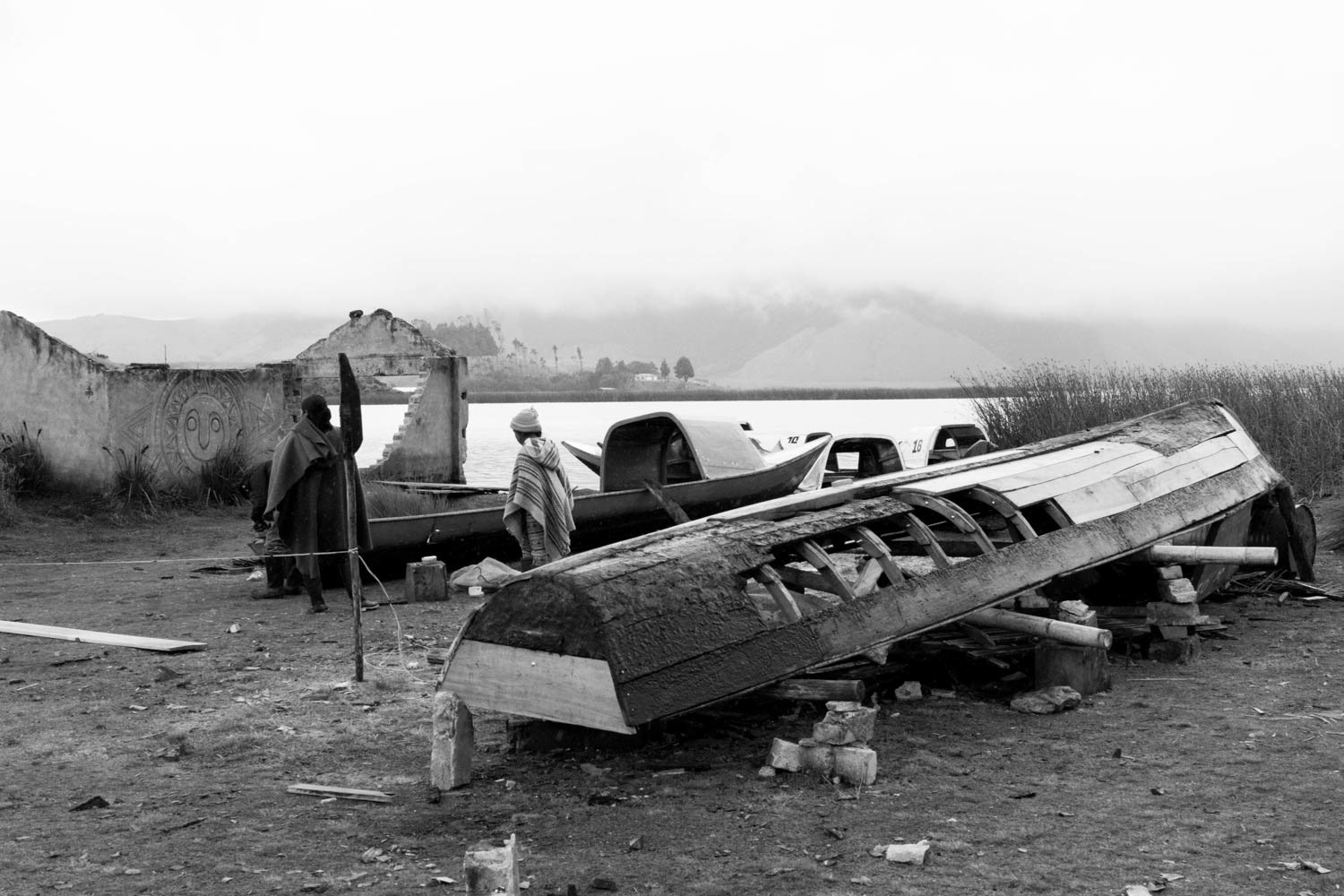 Fotografía en blanco y negro de embarcaciones de madera en reparación junto a lago, con personas y paisaje rural al fondo