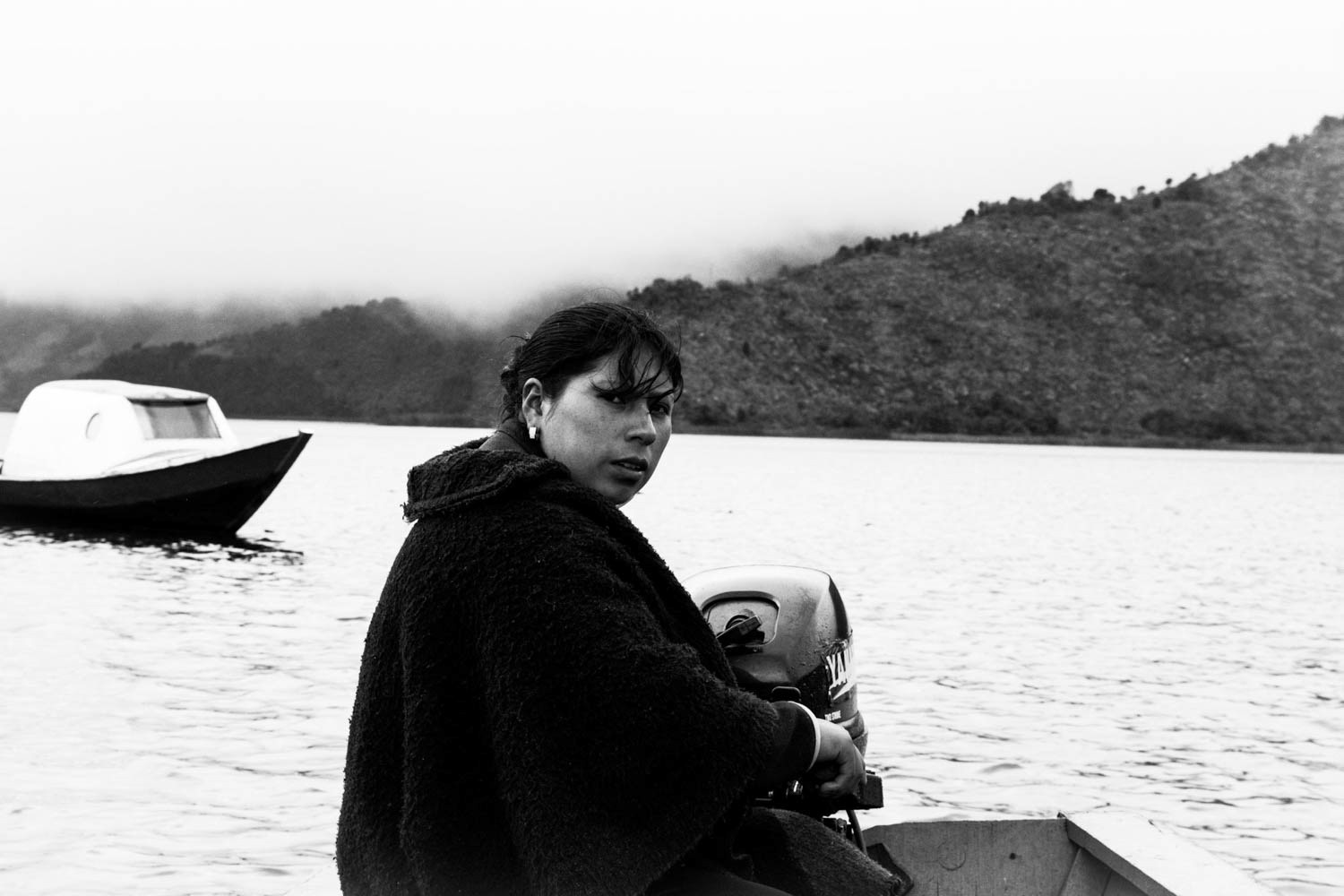 Fotografía en blanco y negro de mujer en bote sobre lago, con montañas cubiertas de niebla y otra embarcación al fondo