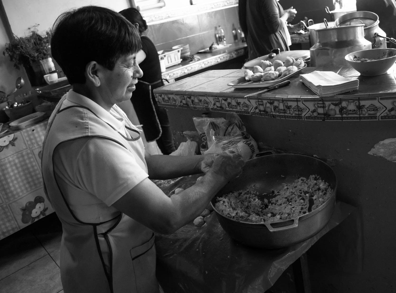 Fotografía en blanco y negro de mujer preparando alimentos en cocina comunitaria, con mesa de trabajo y utensilios tradicionales