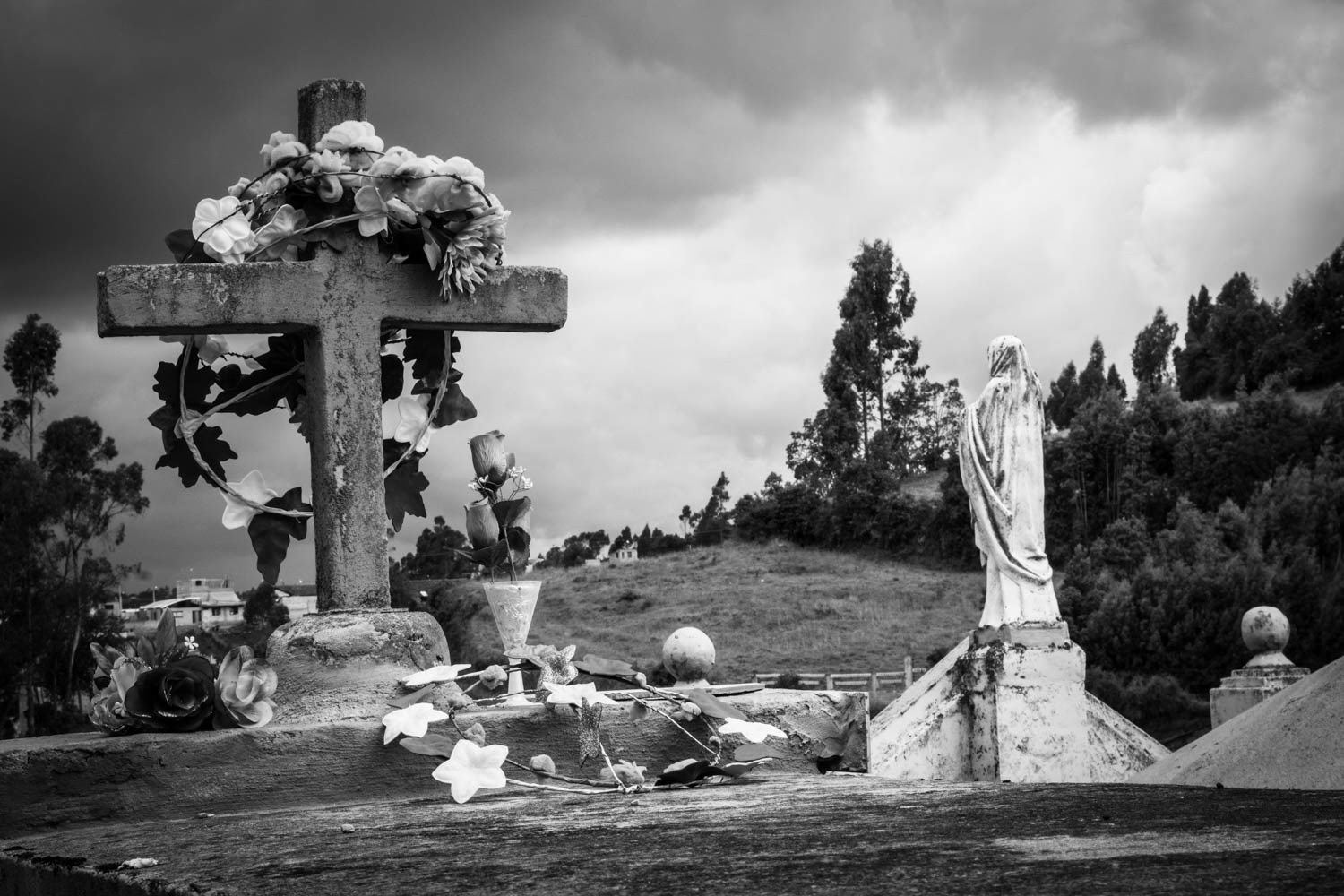 Fotografía en blanco y negro de cruz y estatua religiosa en cementerio rural, con flores y paisaje montañoso al fondo