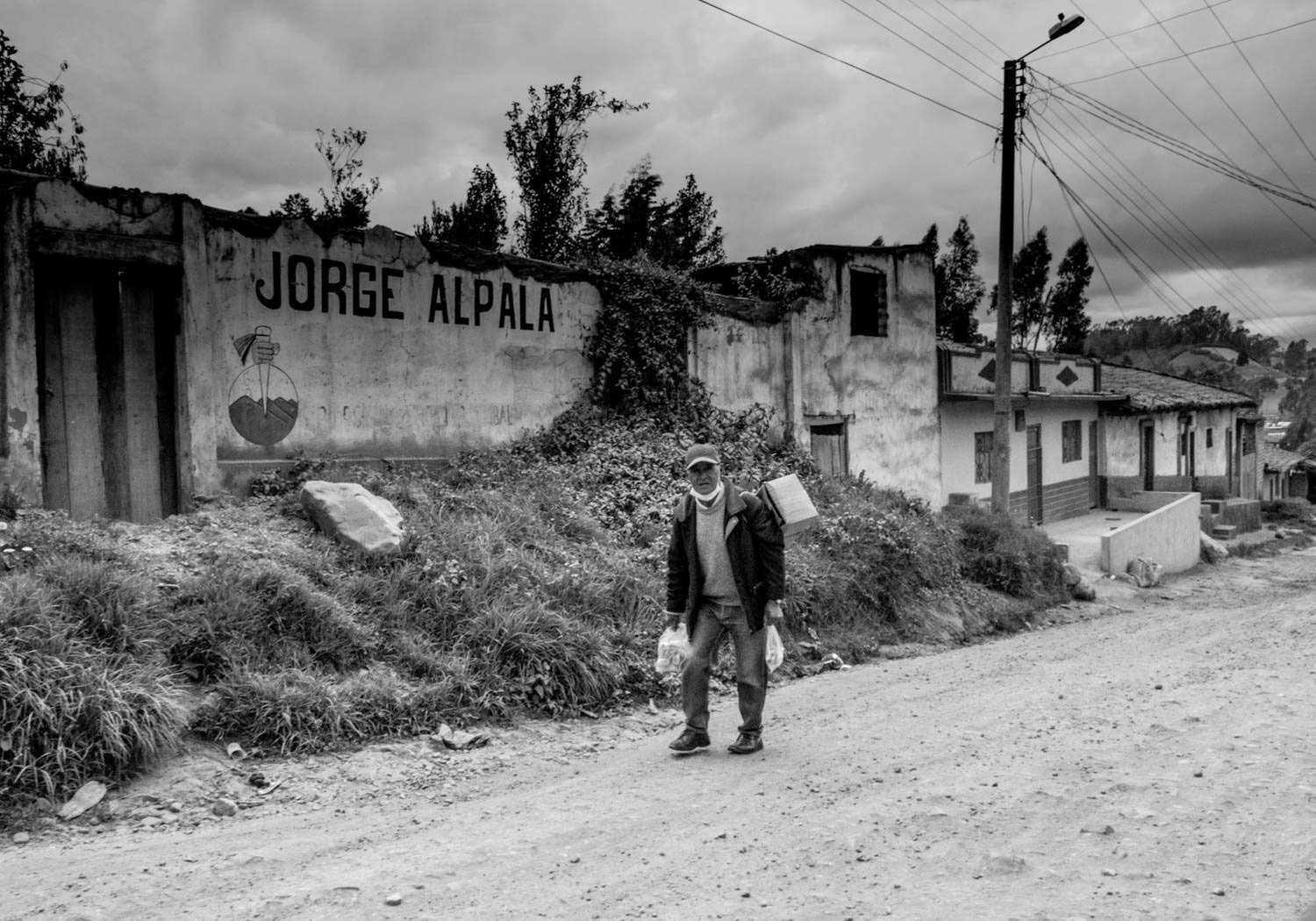Fotografía en blanco y negro de hombre caminando por camino rural frente a muro con grafiti Jorge Alpala y viviendas campesinas
