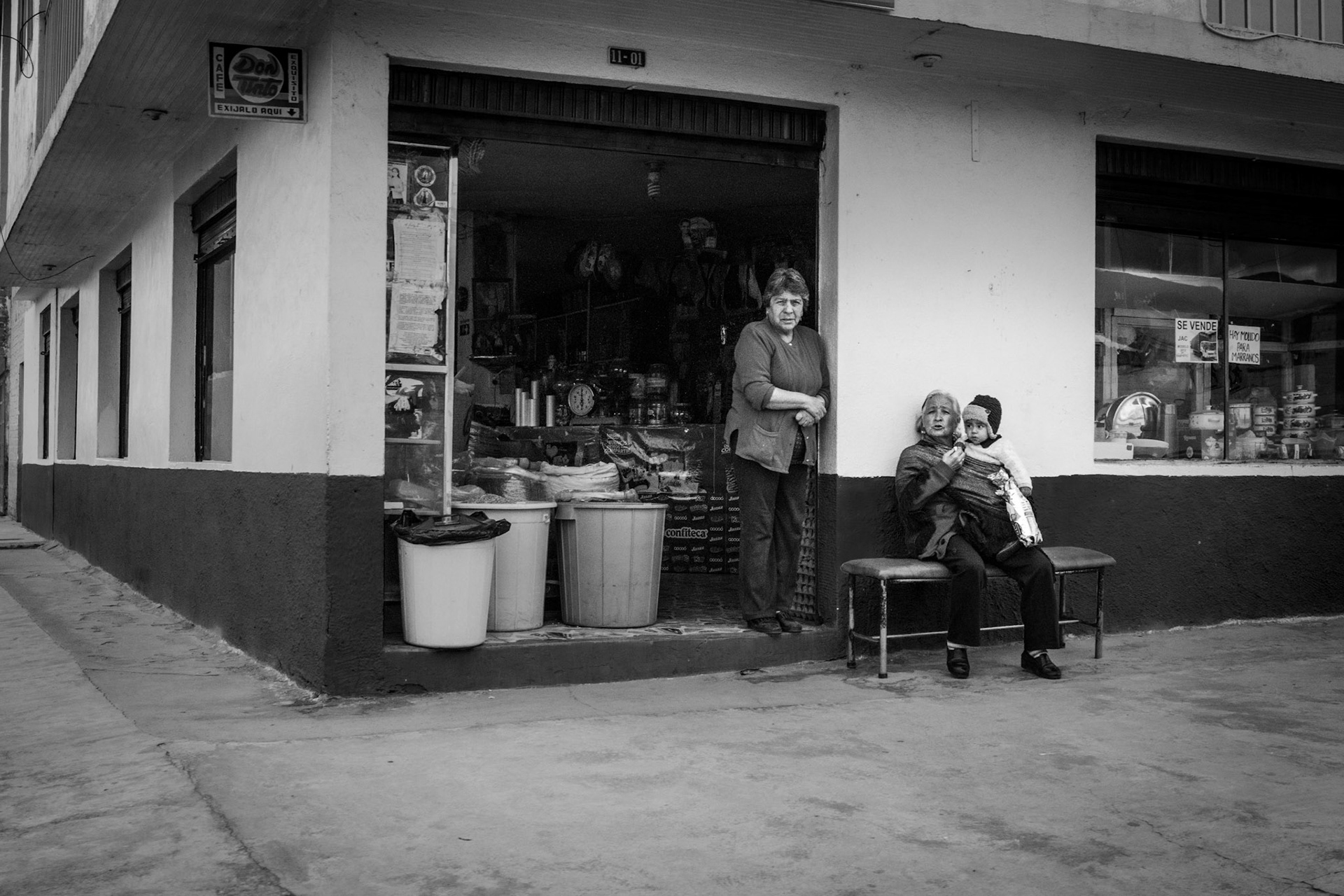 Fotografía en blanco y negro de mujeres frente a tienda de barrio rural, una sentada con niño en brazos y otra en la entrada