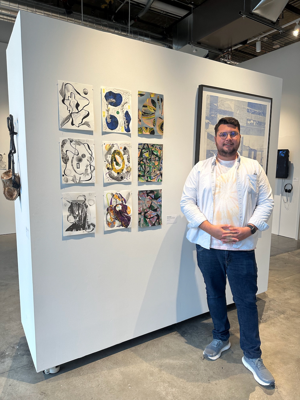 This is a photograph of Daniel Thomas standing by some of his artwork in a gallery. There is a group exhibition with the works of many other artists in the background.