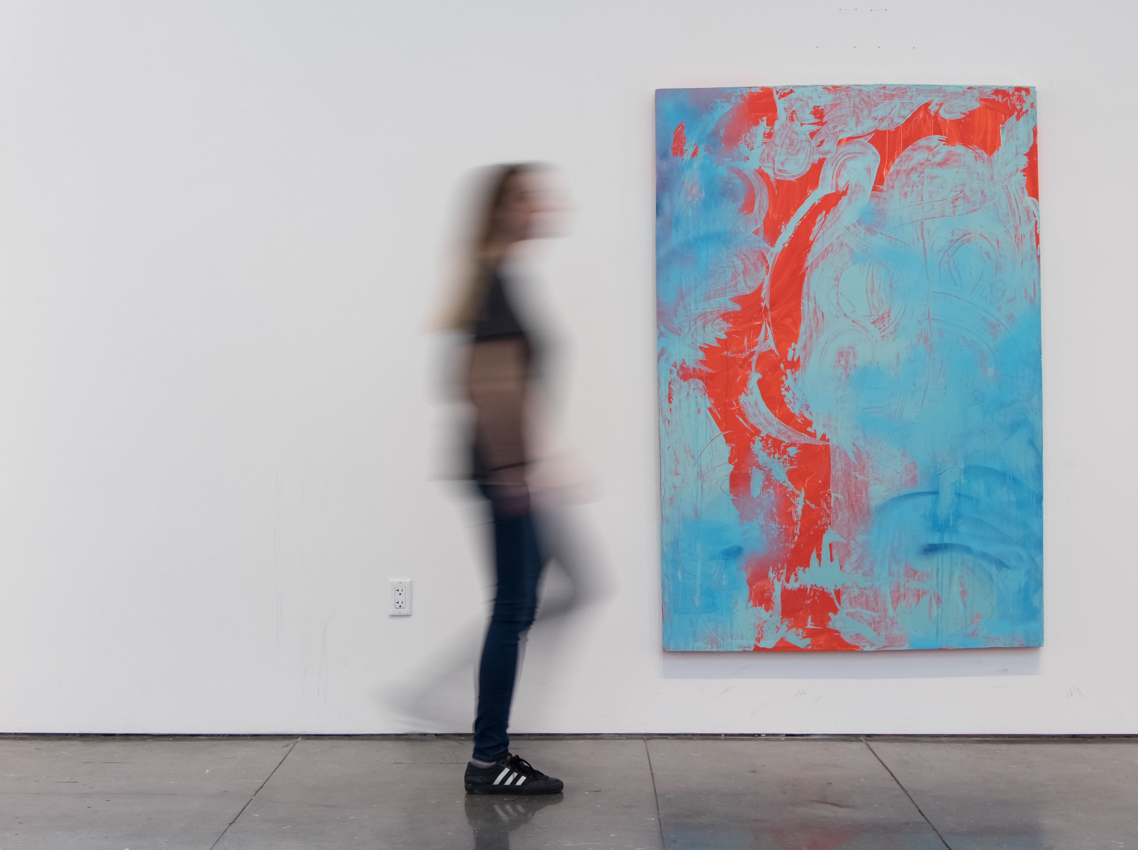 This photograph documents an oversized, abstract painting with a person walking by for scale. The painting is gestural, with an orange background and blue marks made with a scraper and spay paint.