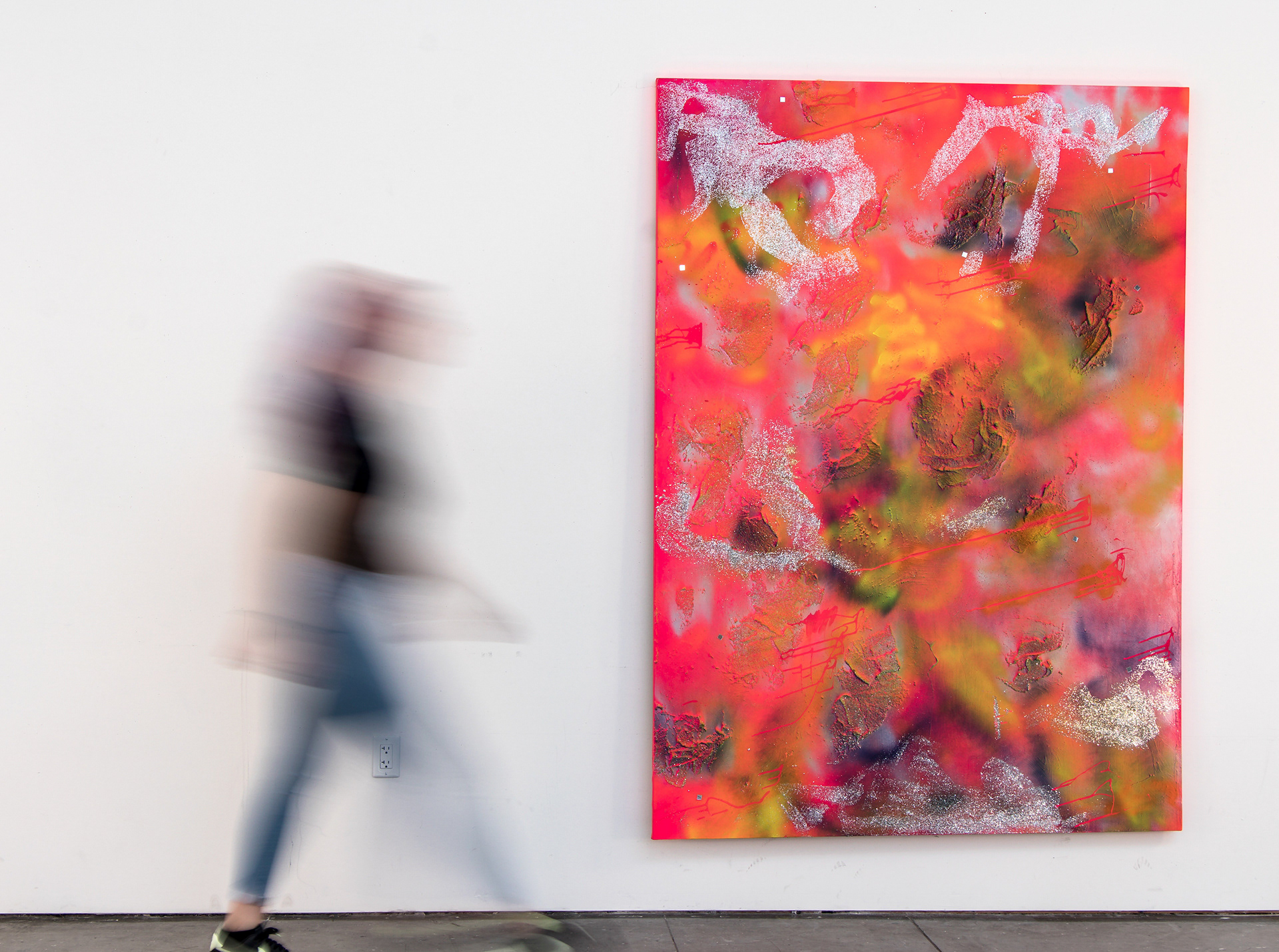 This photograph documents an oversized, abstract painting with a person walking by for scale. The painting is hot pink with blue and yellow areas, glitter, and glass beads.