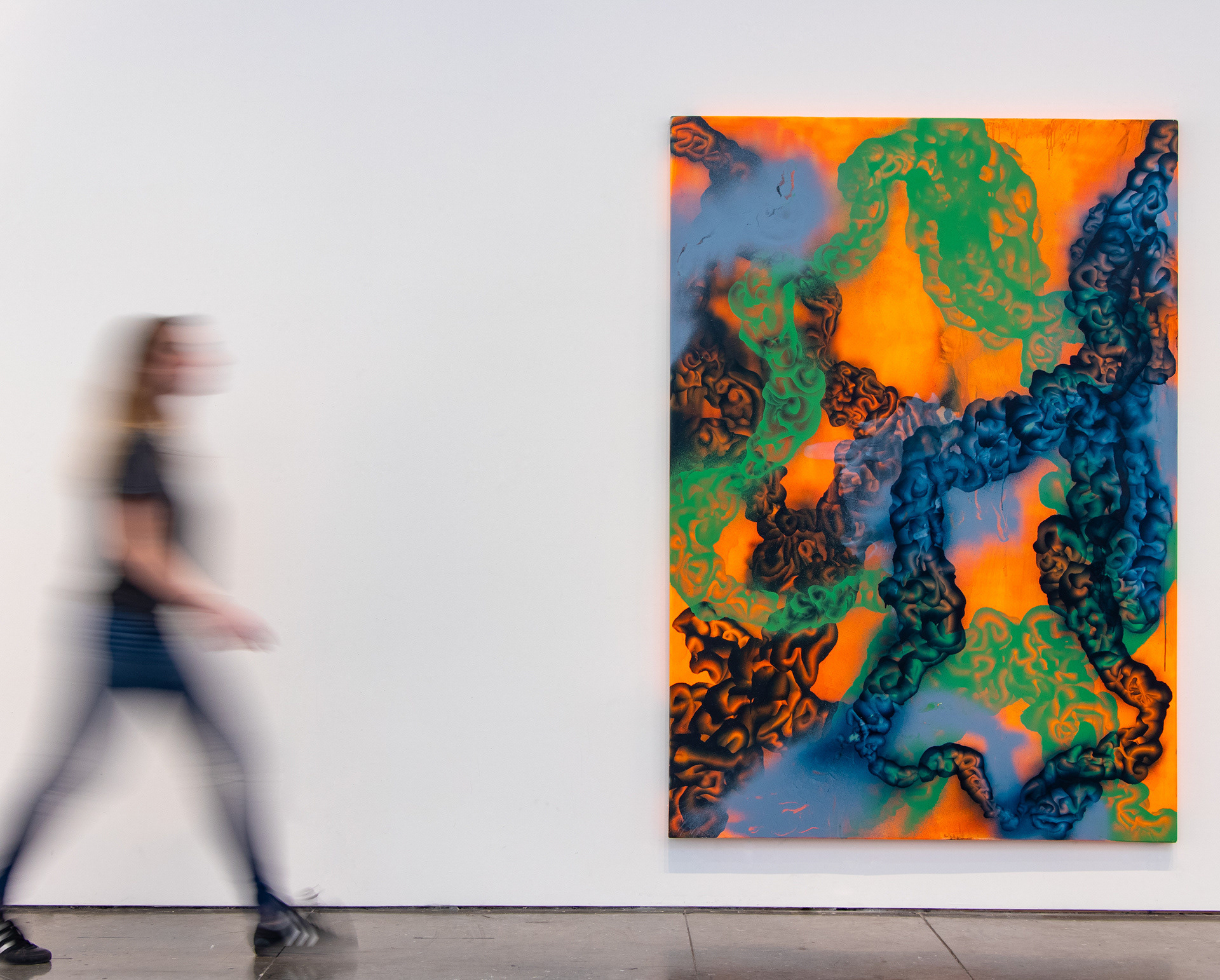 This photograph documents an oversized, abstract painting with a person walking by for scale. The painting features interlocking orange, blue, and green lines.