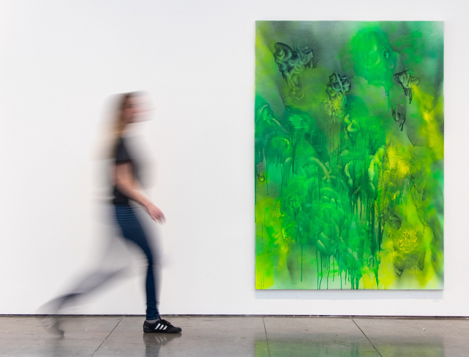 This photograph documents an oversized, abstract painting with a person walking by for scale. The painting is green and gray, composed of irregular blobs and dripping forms.