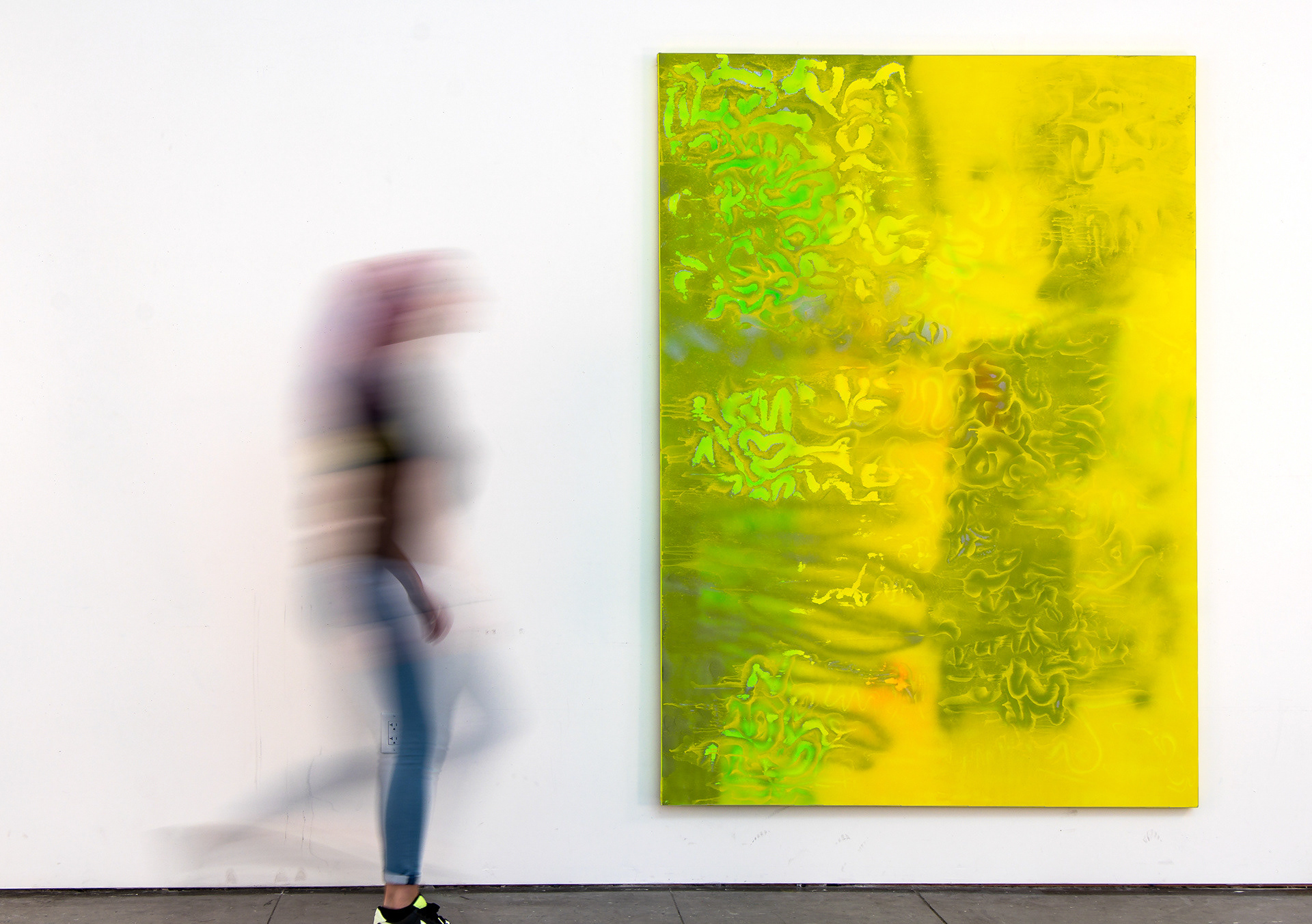 This photograph documents an oversized, abstract painting with a person walking by for scale. The painting features meandering spray paint marks in yellow, green, and gray.