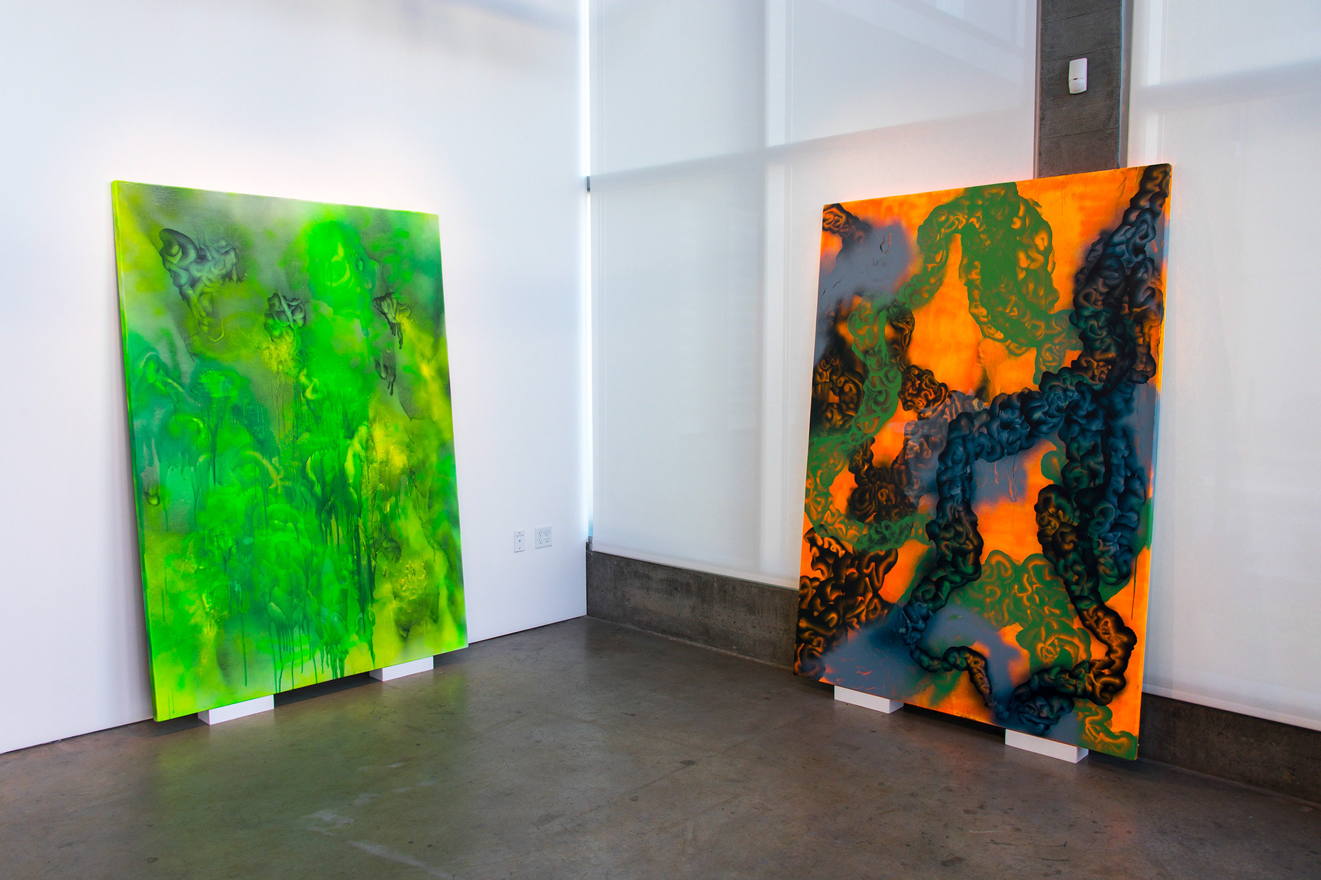 This photograph documents aspects of a painting exhibition called "After Aura." It shows two large paintings, one green, one orange, leaning against a wall and a bank of windows.