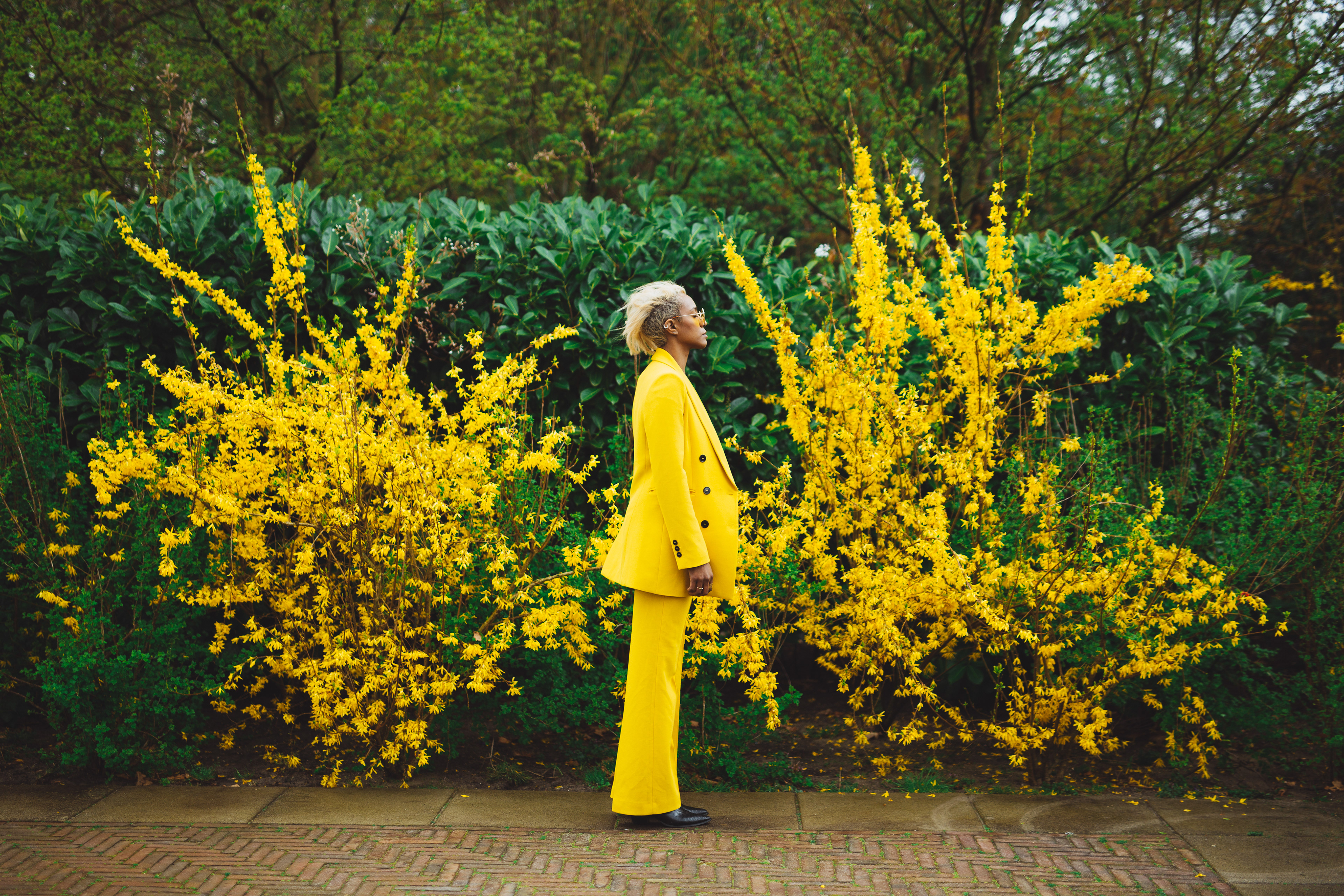 Jasmine in yellow jumpsuit in a profile position posing in front of yellow bushes