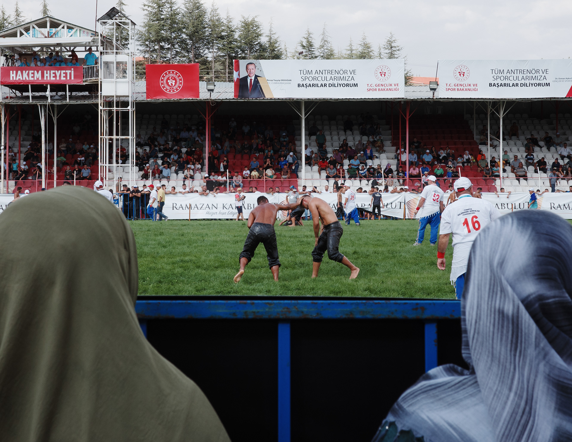 Two women on the sidelines watching two wrestlers duel. 671. Elmalı Oil Wrestling Festival, Türkiye on 8 September 2023.