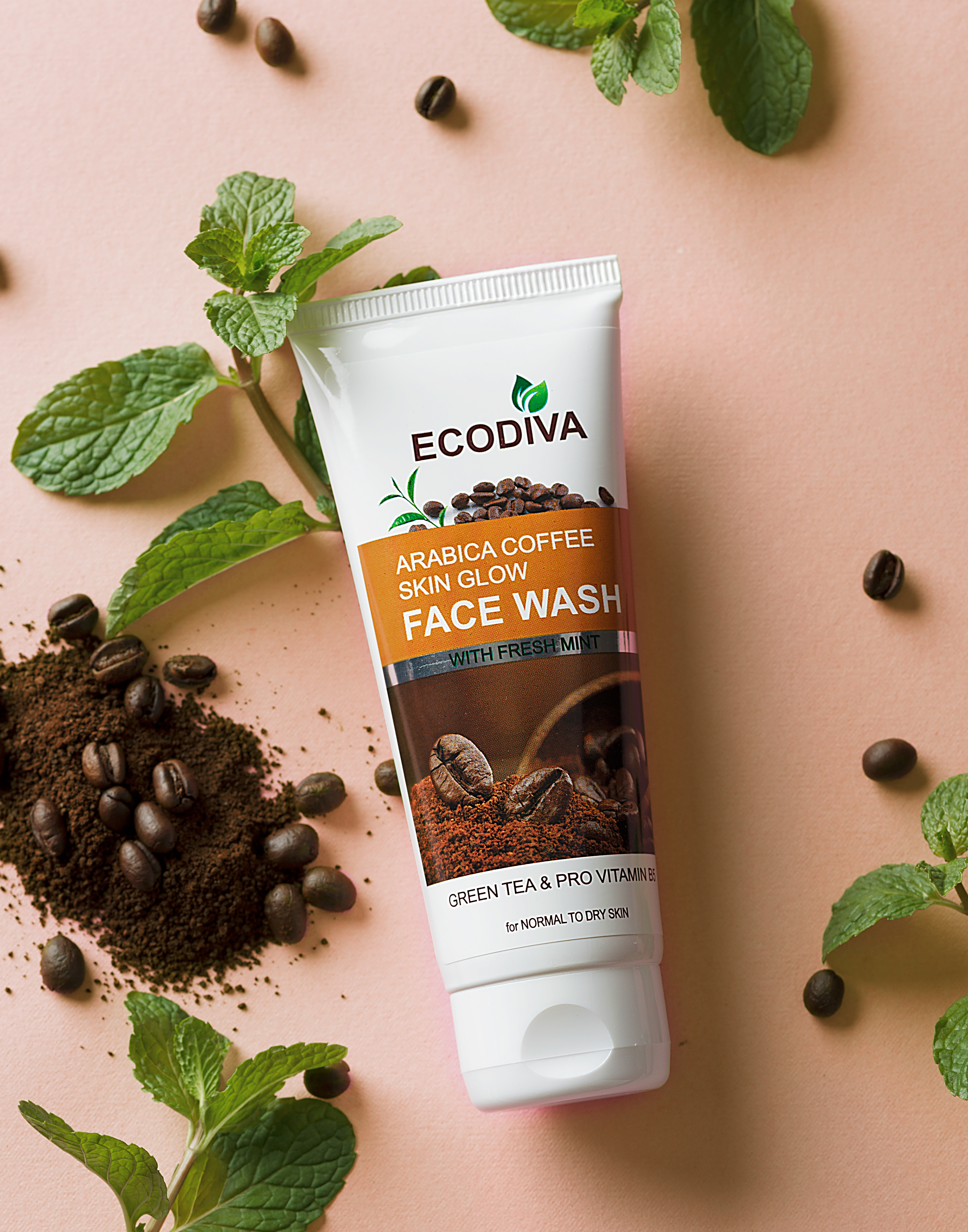 Creative composed image of a tube of Ecodiva Arabica Coffee Skin Glow Face Wash placed on a soft pink background, surrounded by fresh mint leaves and scattered coffee beans. The product packaging highlights key ingredients like green tea, pro-vitamin B5, and fresh mint, designed for normal to dry skin. The rich texture of coffee grounds is visible near the tube, emphasizing the natural, exfoliating properties of the face wash.