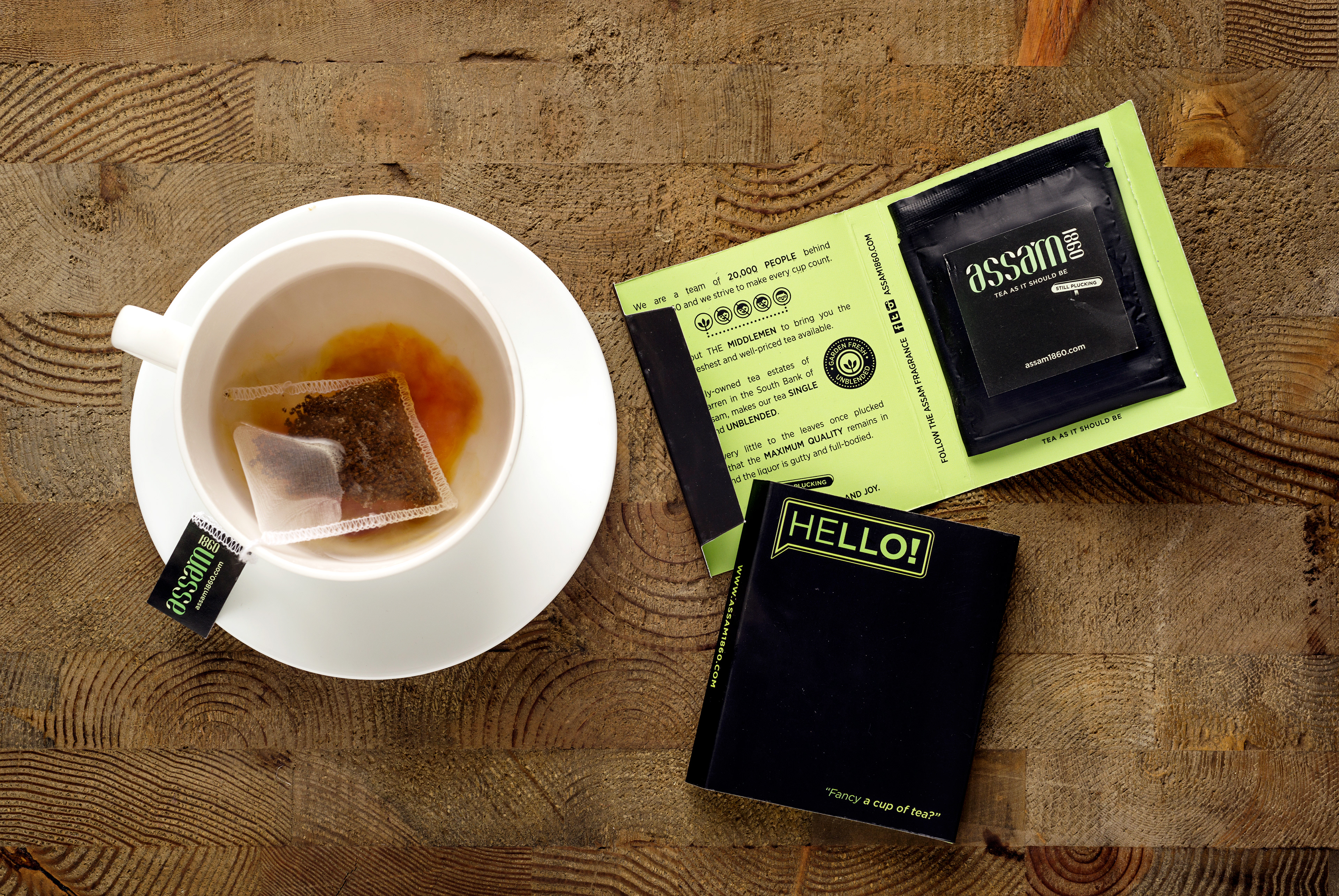 A white cup and saucer on a textured wooden table. Inside the cup, there is a tea bag steeping in hot water, partially infusing the liquid with a brown color. The tea bag label reads "ASSAM TEA," and next to the cup, there is packaging for "Assam 1860" black tea. The packaging is primarily black and green, with a section that says "HELLO!" and "Fancy a cup of tea?" in neon green lettering. The packaging also includes some text explaining the tea's high quality, single-origin nature, and unblended status.