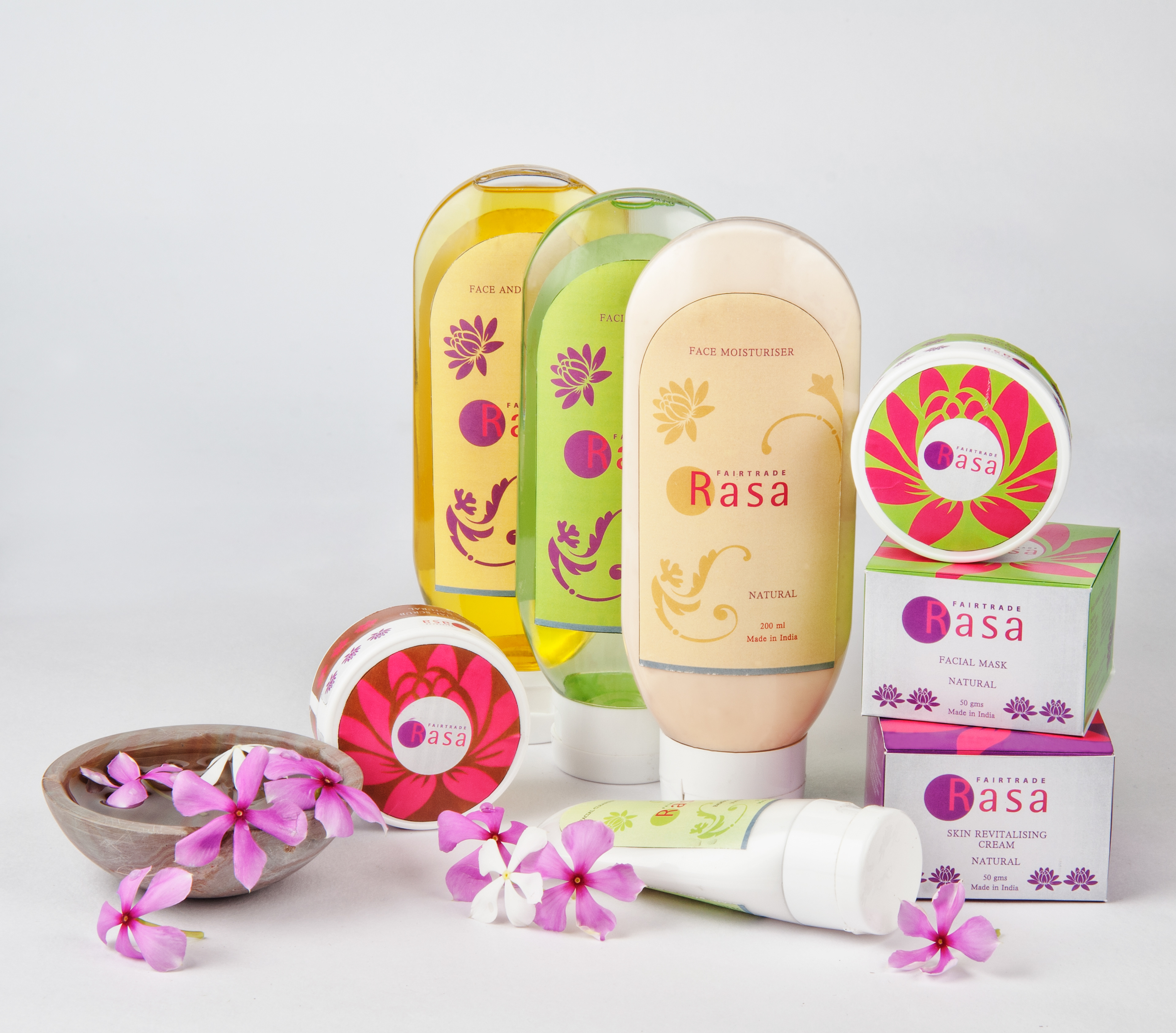 A vibrant display of natural skincare products from the "Rasa" brand. The setup includes various brightly colored containers such as face moisturizer in a gold tube, facial masks in circular tins with pink and green floral designs, and face oils in yellow and green translucent bottles. The products are artfully arranged with pink and white flowers scattered around, enhancing the natural and fresh theme of the skincare line. The packaging prominently states "Fairtrade" and "Made in India," emphasizing natural ingredients and ethical sourcing.