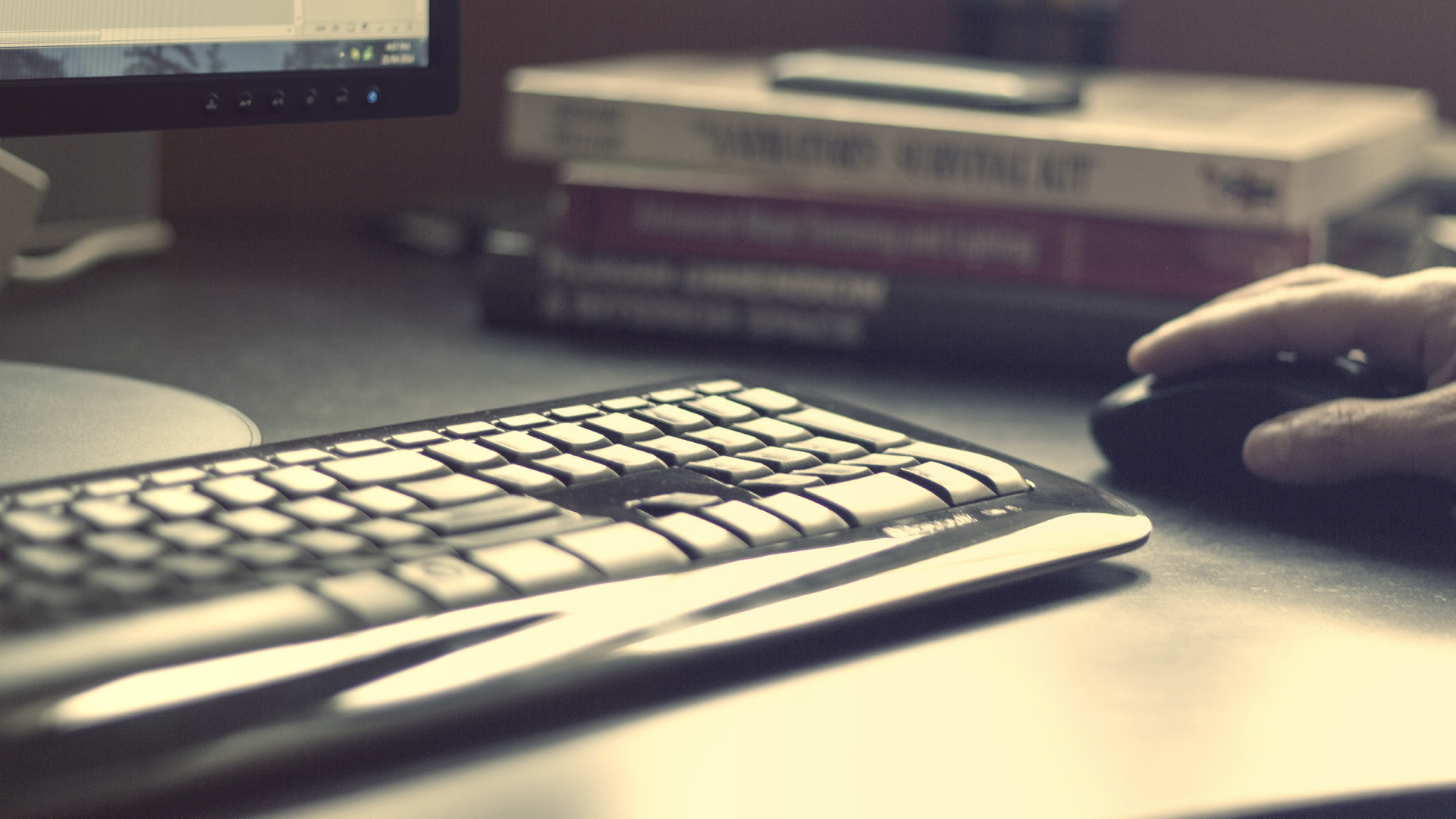 A photograph of a computer desk with a monitor, keyboard, and mouse, with an out-of-focus pile of books in the background.