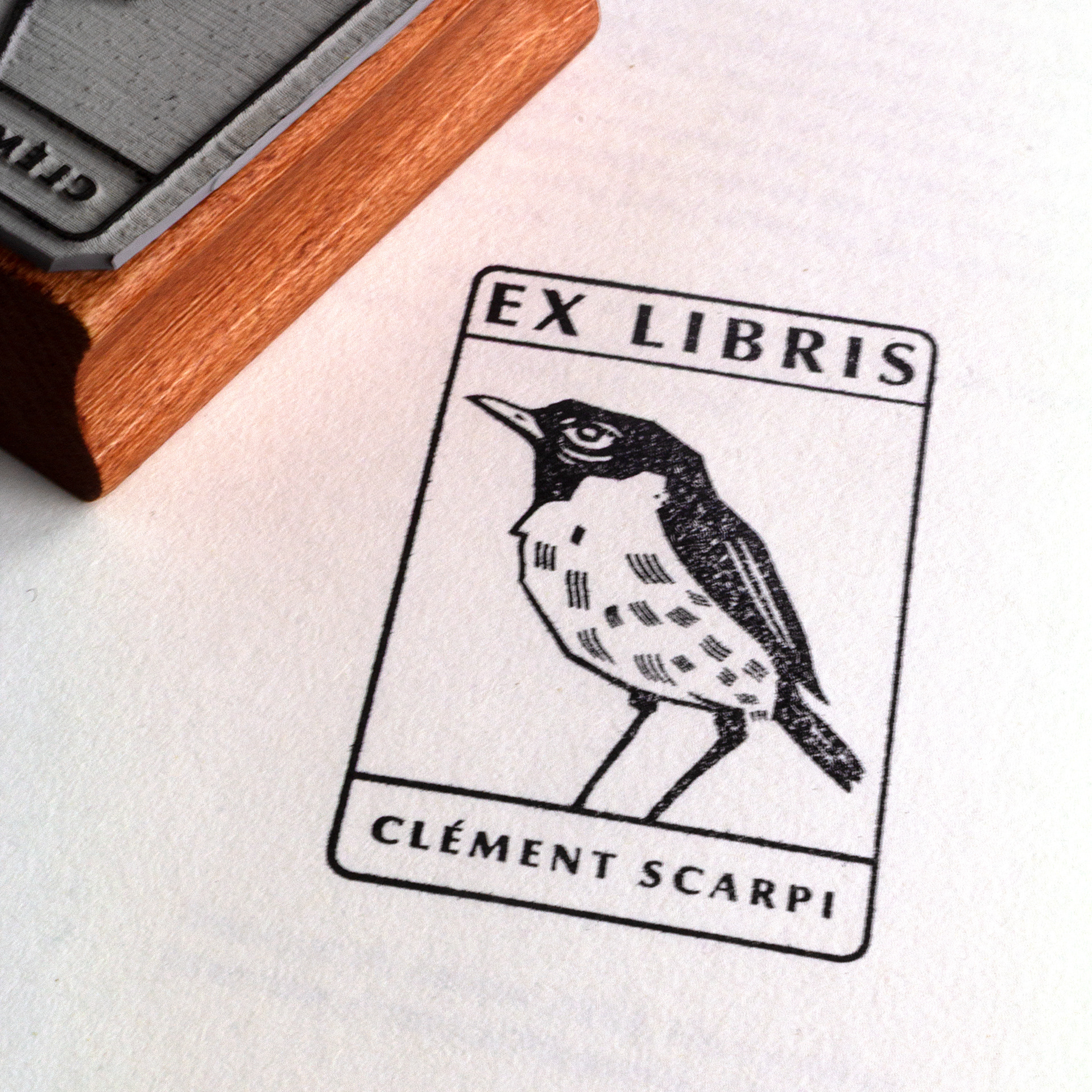 A rubber stamp with a bird illustration and the text “EX LIBRIS CLÉMENT SCARPI” lies next to its stamped impression on white paper.