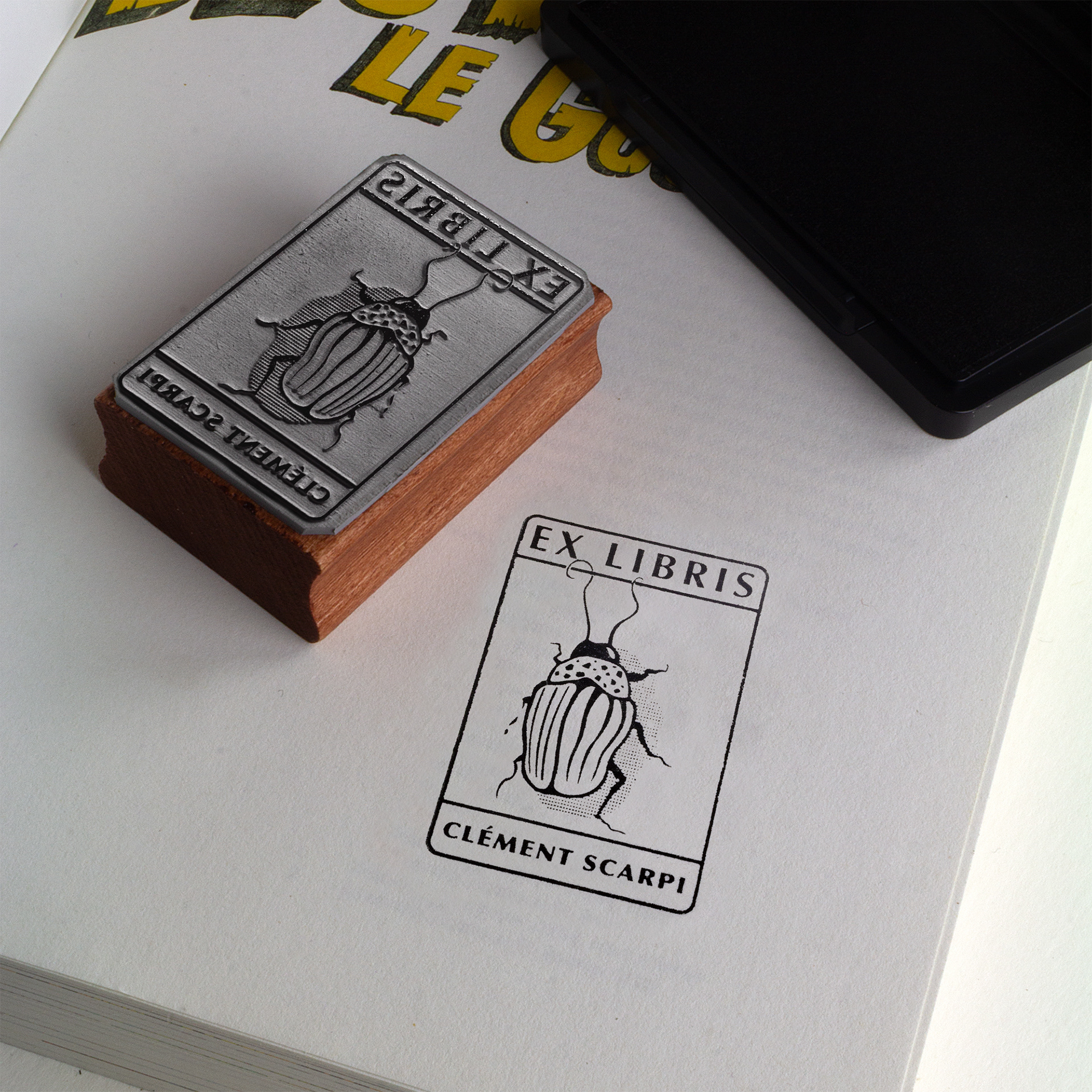 A rubber stamp with a insect illustration and the text “EX LIBRIS CLÉMENT SCARPI” lies next to its stamped impression on white paper.