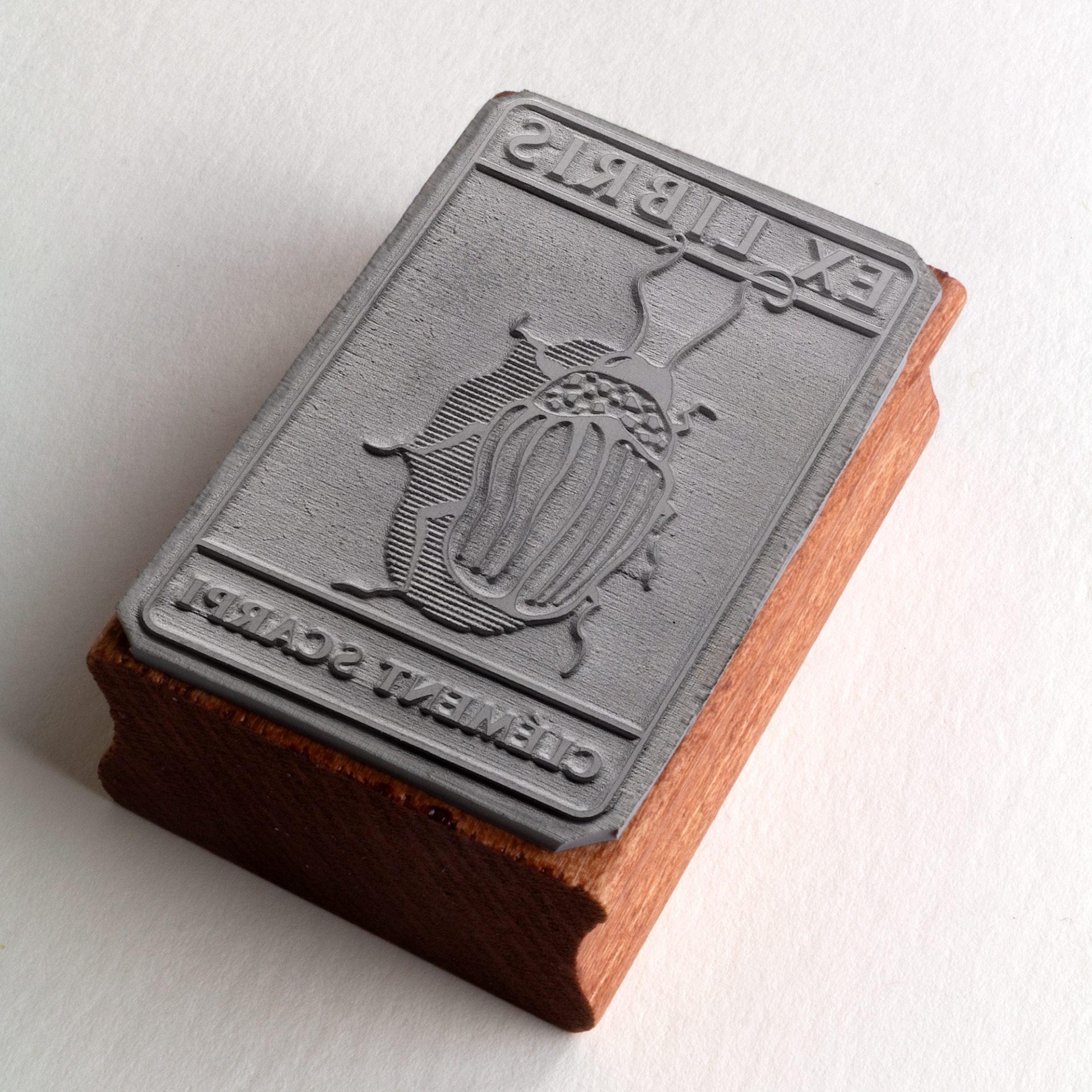 A rubber stamp with a insect illustration and the text “EX LIBRIS CLÉMENT SCARPI” lies next to its stamped impression on white paper.