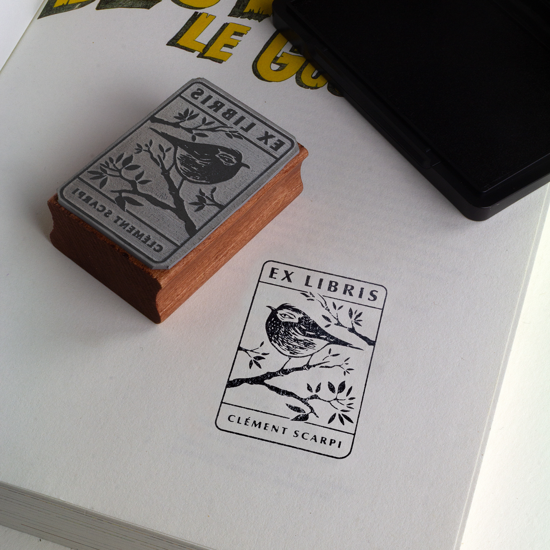 A book with an Ex Libris stamp showing a bird on a branch, the name Clément Scarpi, the wooden stamp, and an ink pad placed nearby