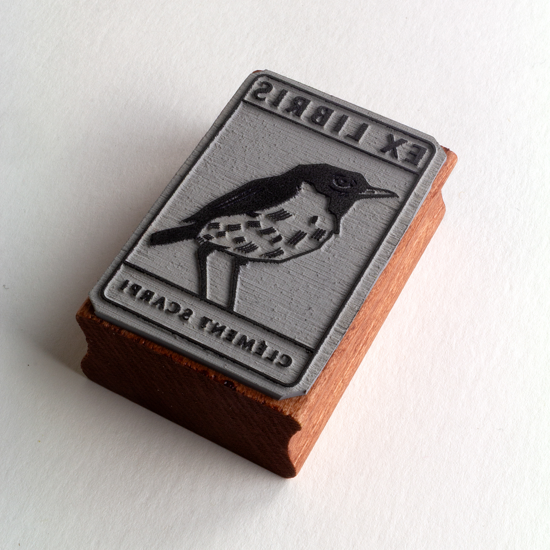 A rubber stamp with a bird illustration and the text “EX LIBRIS CLÉMENT SCARPI” lies next to its stamped impression on white paper.