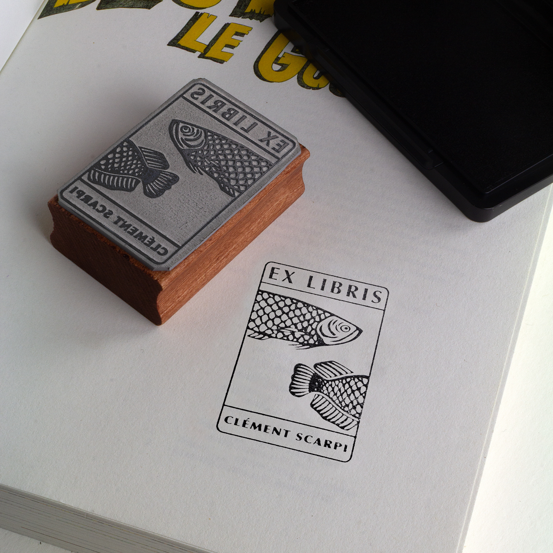 A bookplate stamp with fish illustrations and the words EX LIBRIS CLÉMENT SCARPI is shown next to its inked impression on a white book cover, perfect for showcasing on your personalized website or Adobe Portfolio. An ink pad is partially visible in the background.