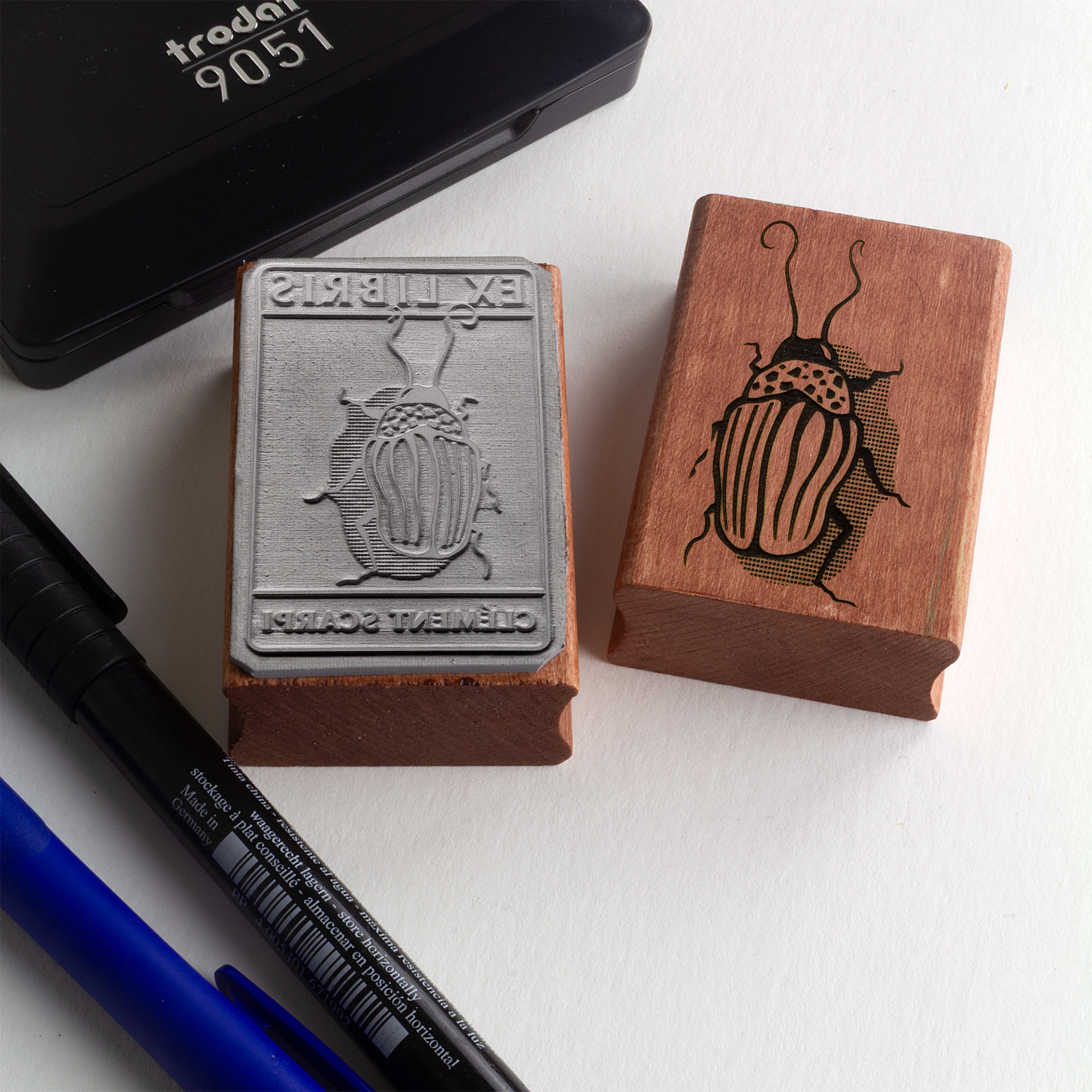 A rubber stamp with a insect illustration and the text “EX LIBRIS CLÉMENT SCARPI” lies next to its stamped impression on white paper.