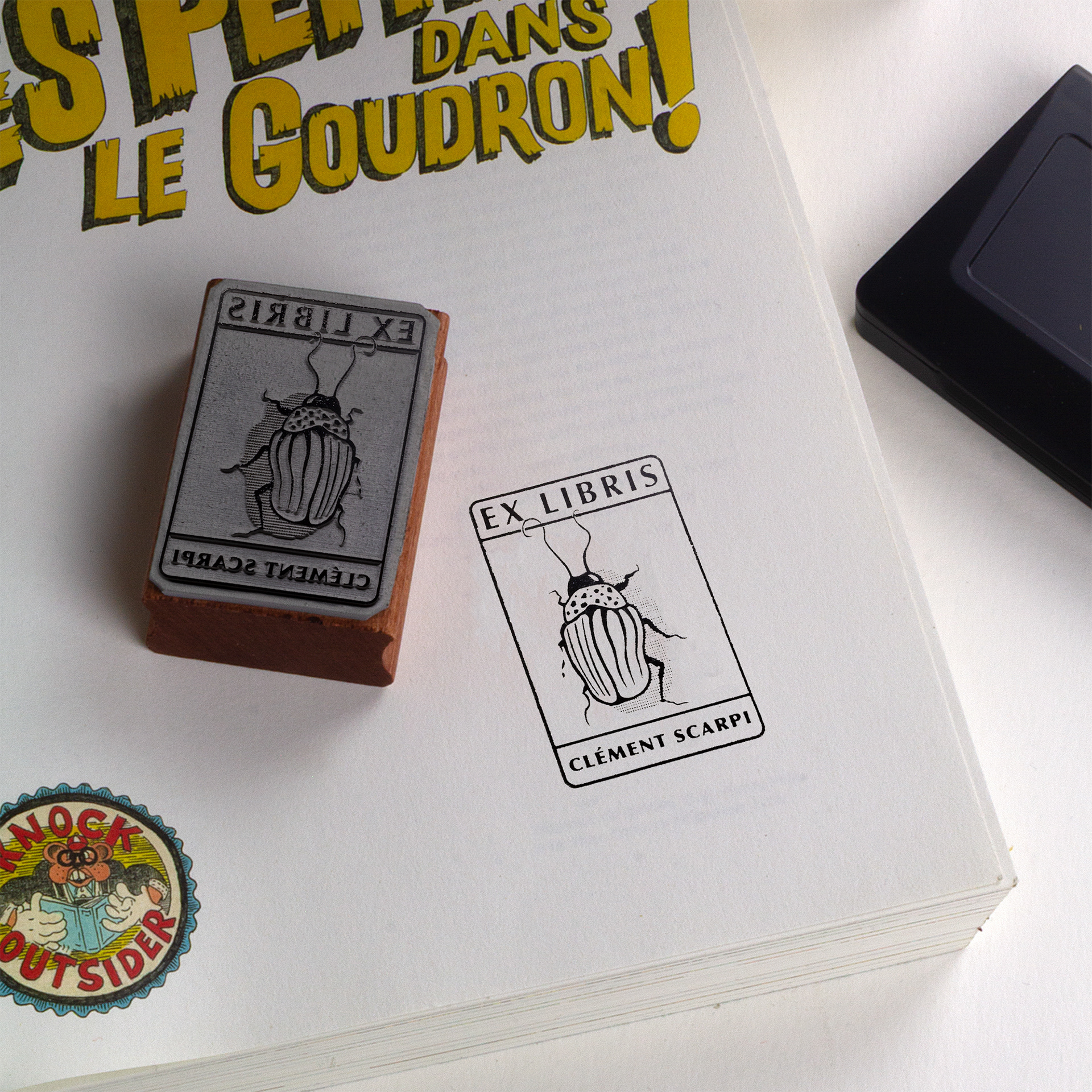 A rubber stamp with a insect illustration and the text “EX LIBRIS CLÉMENT SCARPI” lies next to its stamped impression on white paper.
