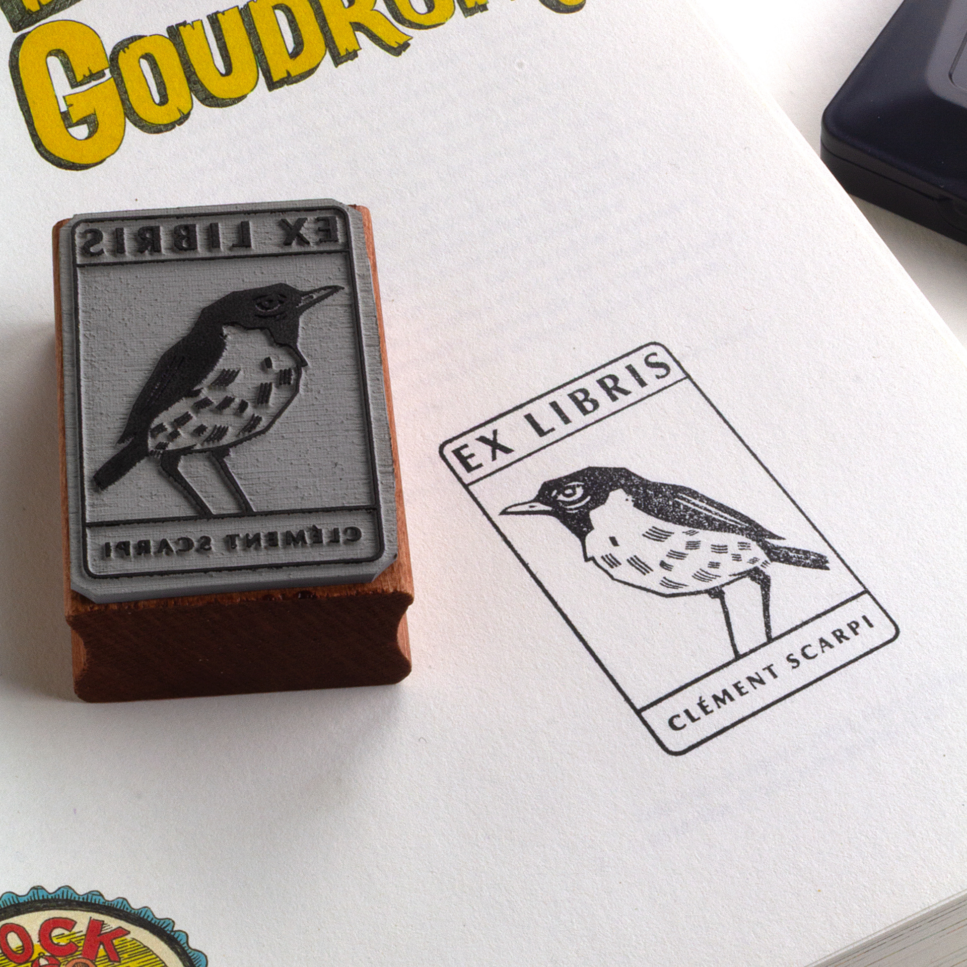 A rubber stamp with a bird illustration and the text “EX LIBRIS CLÉMENT SCARPI” lies next to its stamped impression on white paper.