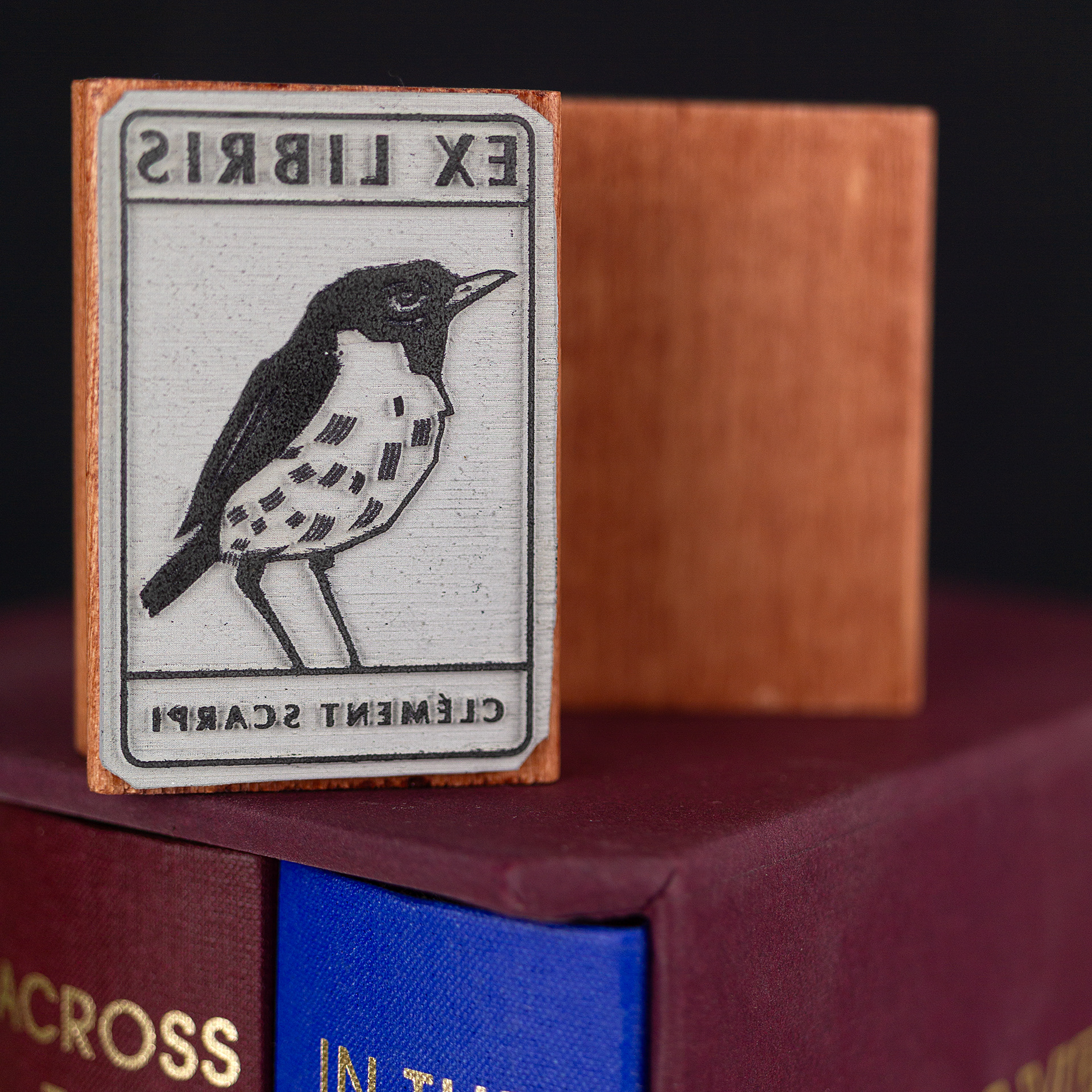 A rubber stamp with a bird illustration and the text “EX LIBRIS CLÉMENT SCARPI” lies next to its stamped impression on white paper.