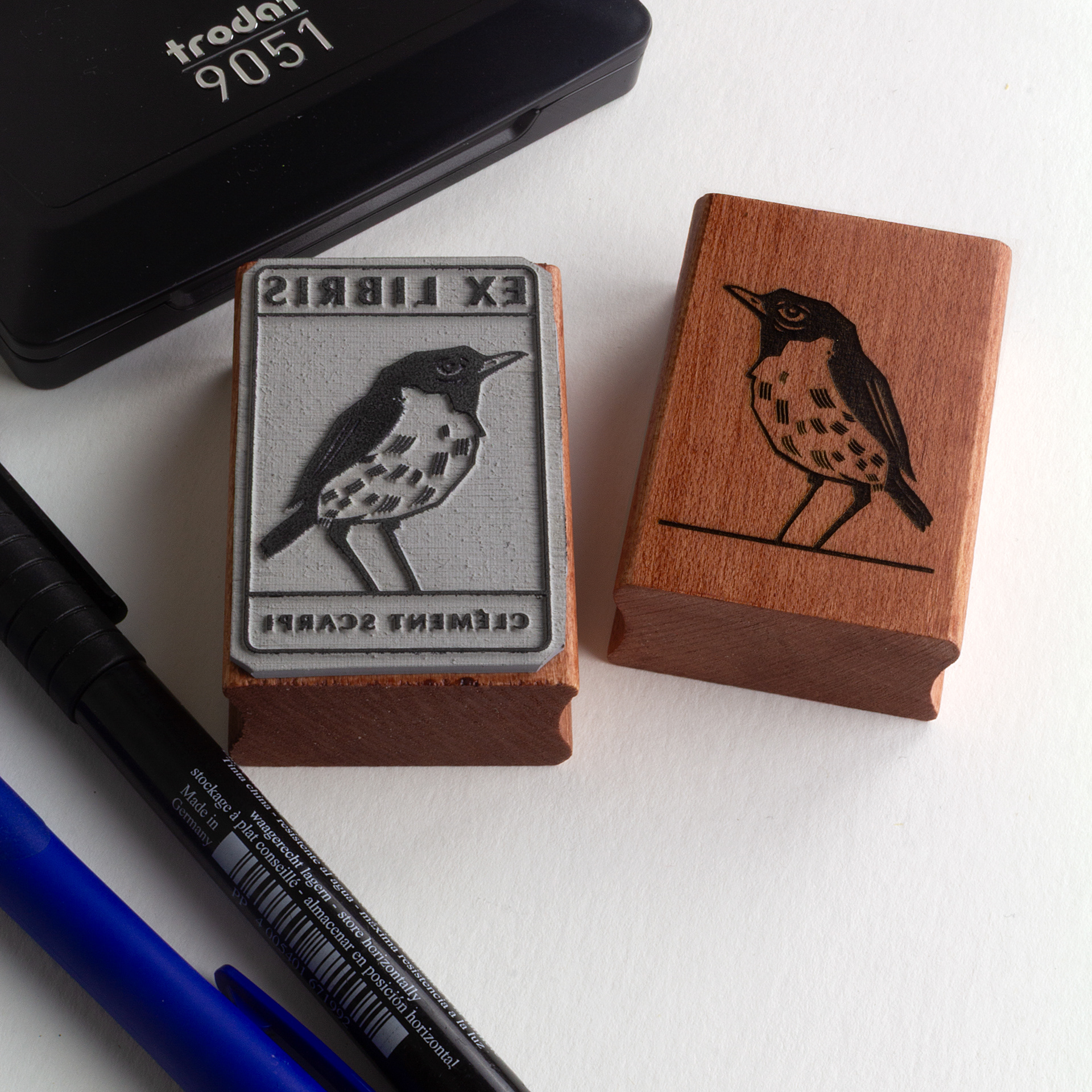 A rubber stamp with a bird illustration and the text “EX LIBRIS CLÉMENT SCARPI” lies next to its stamped impression on white paper.