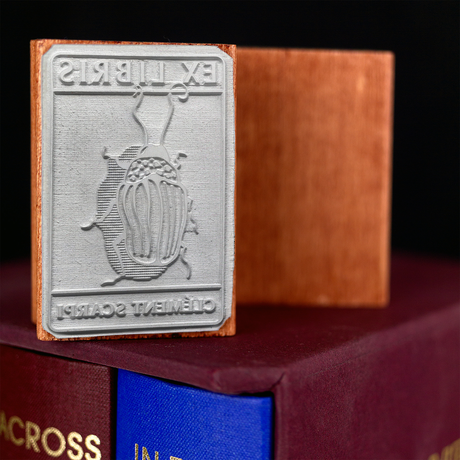 A rubber stamp with a insect illustration and the text “EX LIBRIS CLÉMENT SCARPI” lies next to its stamped impression on white paper.