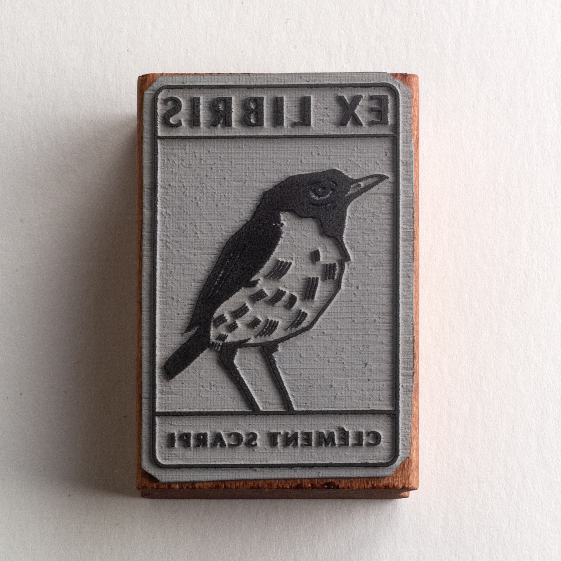A rubber stamp with a bird illustration and the text “EX LIBRIS CLÉMENT SCARPI” lies next to its stamped impression on white paper.