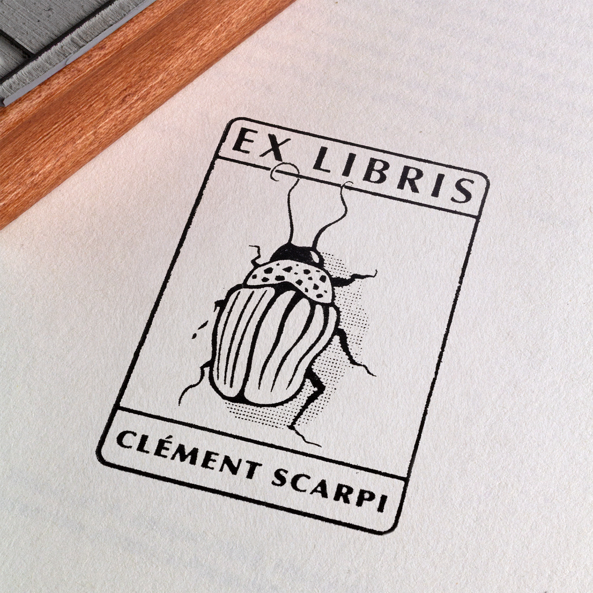 A rubber stamp with a insect illustration and the text “EX LIBRIS CLÉMENT SCARPI” lies next to its stamped impression on white paper.