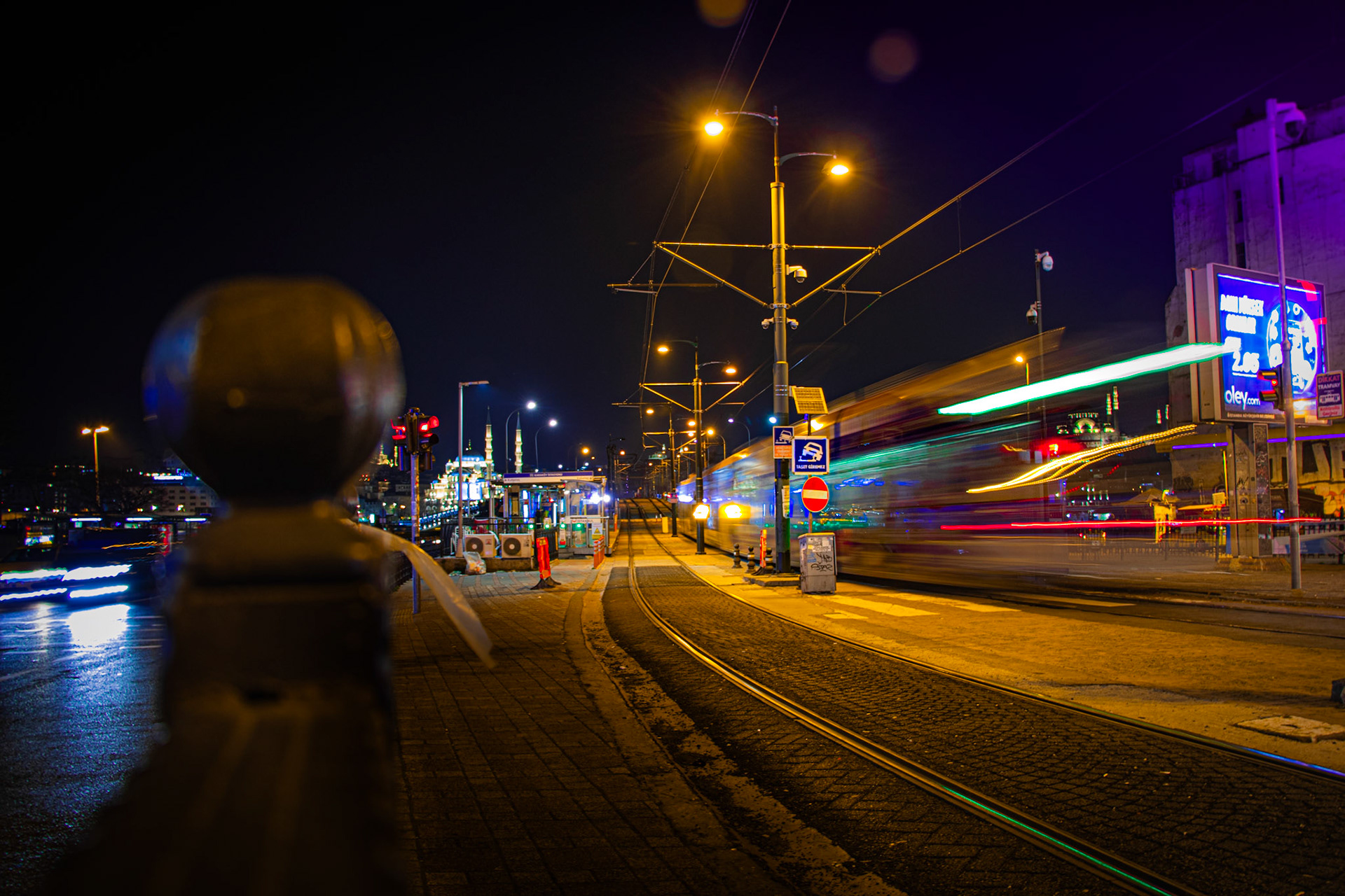Galata Köprüsü'nün ışıl ışıl atmosferinde, gecenin karanlığını yırtan bir tramvay, istasyona yanaşıyor.  Tramvayın ışıkları ve köprünün ihtişamı adeta bir tabloyu andırıyor. Hareket ve durağanlığın bir araya geldiği bu kare, şehrin gizemli ve büyüleyici ruhunu yansıtıyor.