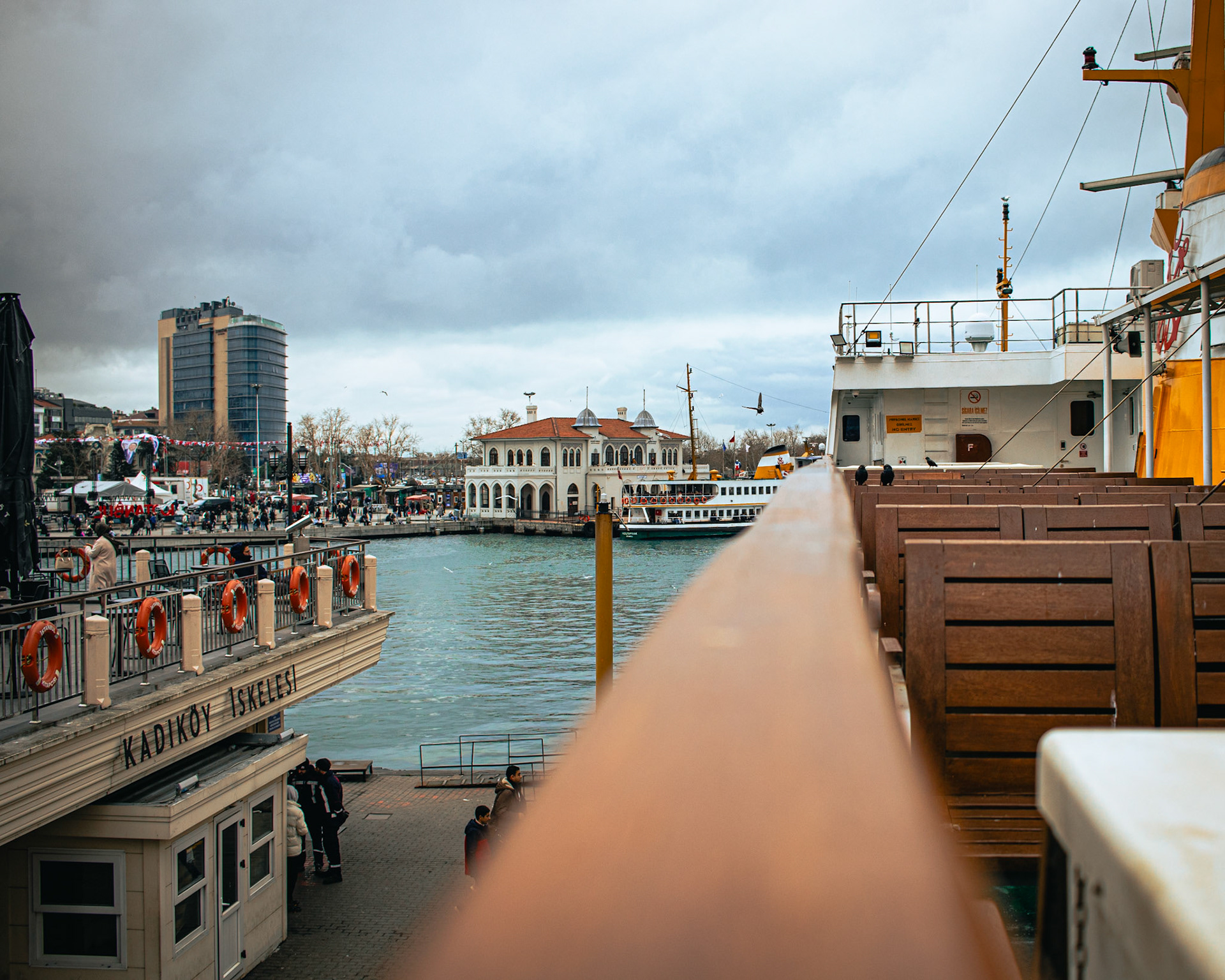 Fotoğrafın yarısını bomboş Kadıköy İskelesi, diğer yarısını ise güvertedeki koltuklar oluşturuyor. Koltukların üzerinde ise yan yana duran iki karga, sessiz bir sohbete dalmış gibi görünüyor. Bu manzara, şehrin karmaşasından uzak bir huzur köşesi sunuyor ve adeta bir meditasyona davet ediyor. Karganın siyah tüyleri ve iskelenin gri tonları, fotoğrafın melankolik atmosferine katkıda bulunuyor.