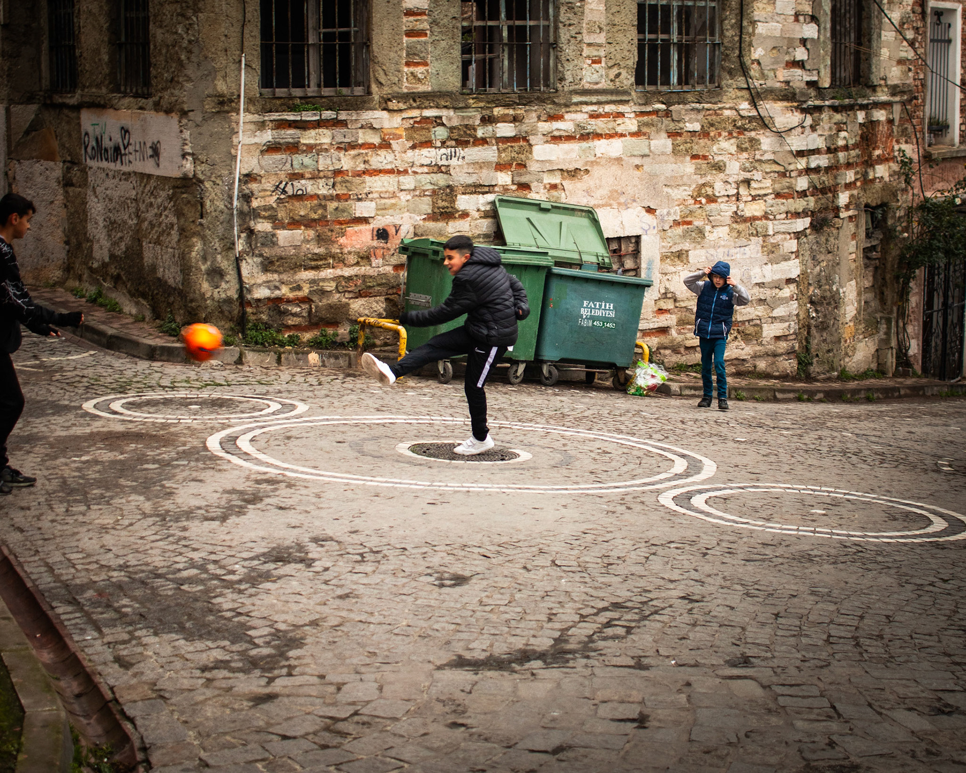 Tarihi bir sokakta, arnavut kaldırımlı zemin üzerinde çocuklar turuncu bir topla oynuyorlar. Çocukların neşeli kahkahaları ve topun sekme sesi tarihi sokakta yankılanıyor. Turuncu top, bir renk cümbüşü gibi sokakta adeta dans ediyor.