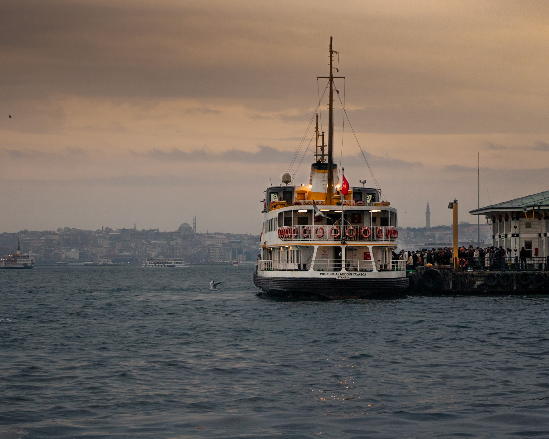 Gün batımının altın rengi ışıkları denizin üzerinde dans ederken, iskeleye yanaşan vapur, yeni bir hikayeye yelken açıyor. Binen yolcuların yüzlerinde farklı duygular var. Kimisi eve dönmenin huzurunu yaşıyor, kimisi yeni bir maceraya atılmanın heyecanını. Vapurun düdüğü öterek ayrılırken, martılar da bu vedaya eşlik ediyor. Güvertede, şehrin silueti ve batmakta olan güneşin muhteşem manzarası karşısında büyülenen yolcular, sessiz bir hayranlıkla bu anı yaşıyor.
