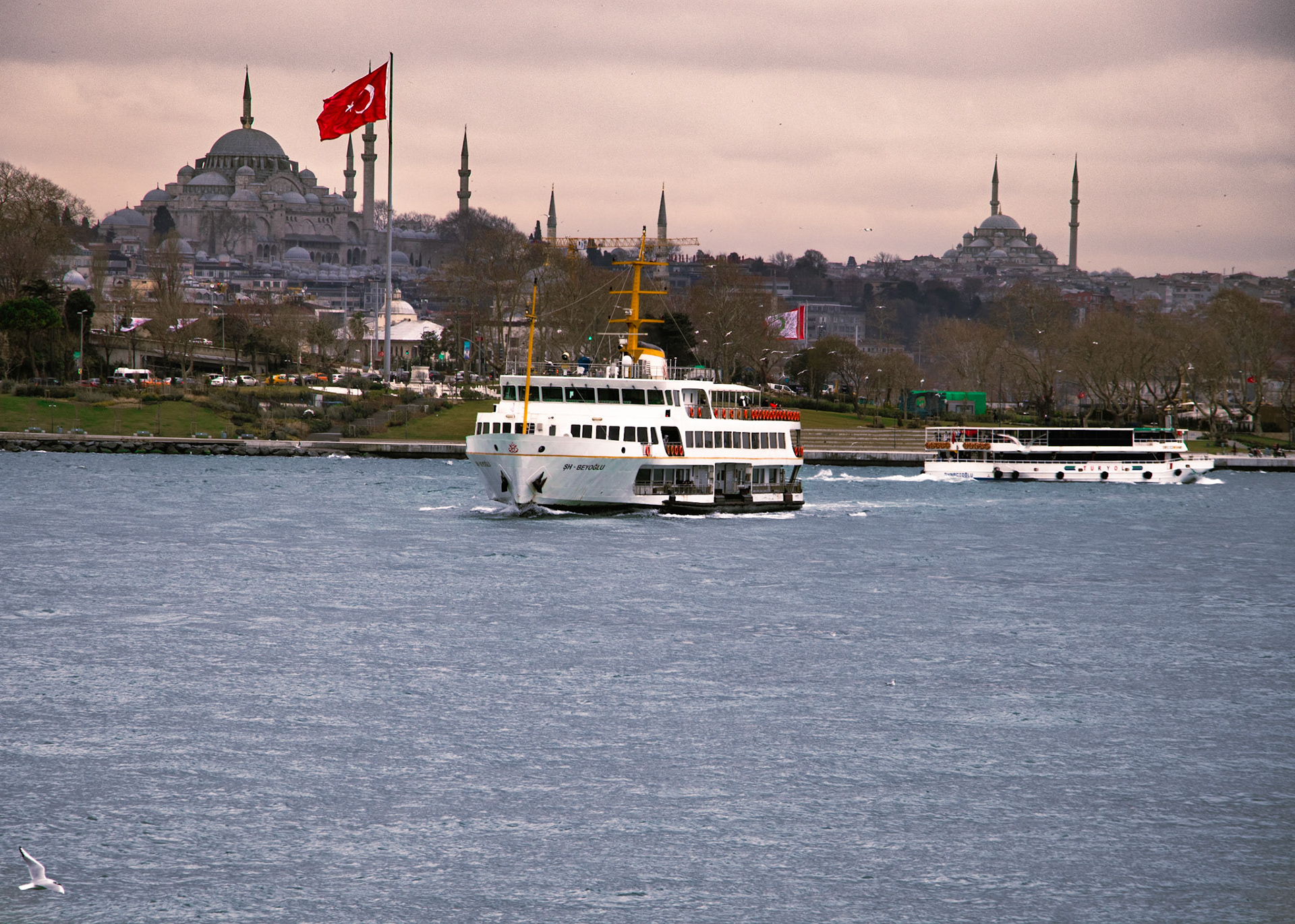 Tarihi Yarımada'dan ayrılan ŞH-Beyoğlu vapuru, Sultanahmet Camii ve Şanlı Türk Bayrağı'nın heybetli manzarasının önünden geçiyor. Bu kare, adeta bir tabloyu andırıyor. Bir yanda tarihi caminin ihtişamı, diğer yanda gururla dalgalanan bayrağımız, vatan sevgisini ve tarihi bilinci körüklüyor. Vapurun denizin üzerinde bıraktığı izler ise adeta bir veda mesajı gibi, geride kalan tarihi yarımadaya selam veriyor.