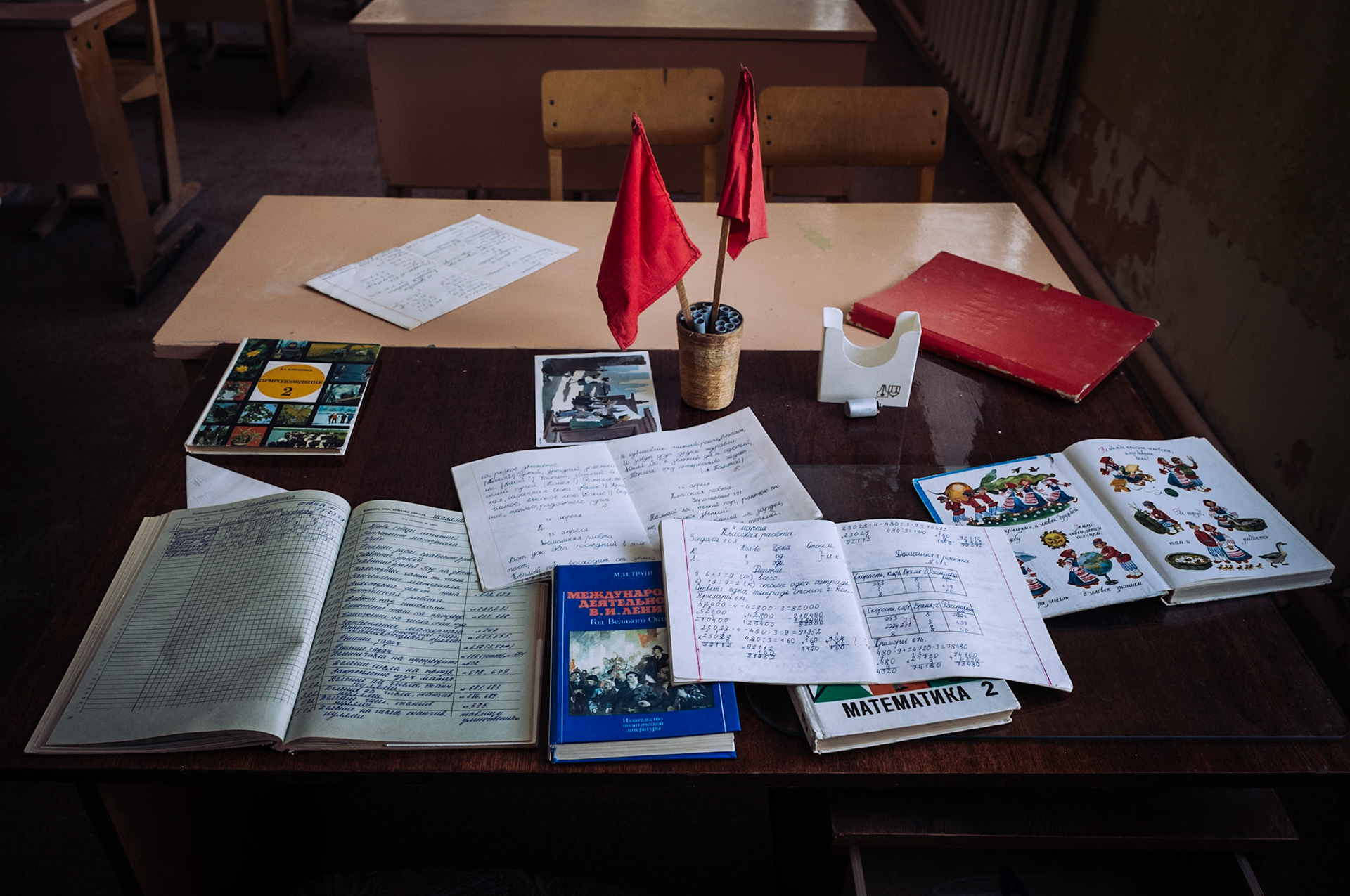 Ein Lehrerpult mit offenen Schulheften, Büchern und zwei kleinen roten Fahnen in der verlassenen Schule von Pyramiden. Die Schriften und Lehrmaterialien spiegeln die sowjetische Erziehung wider.