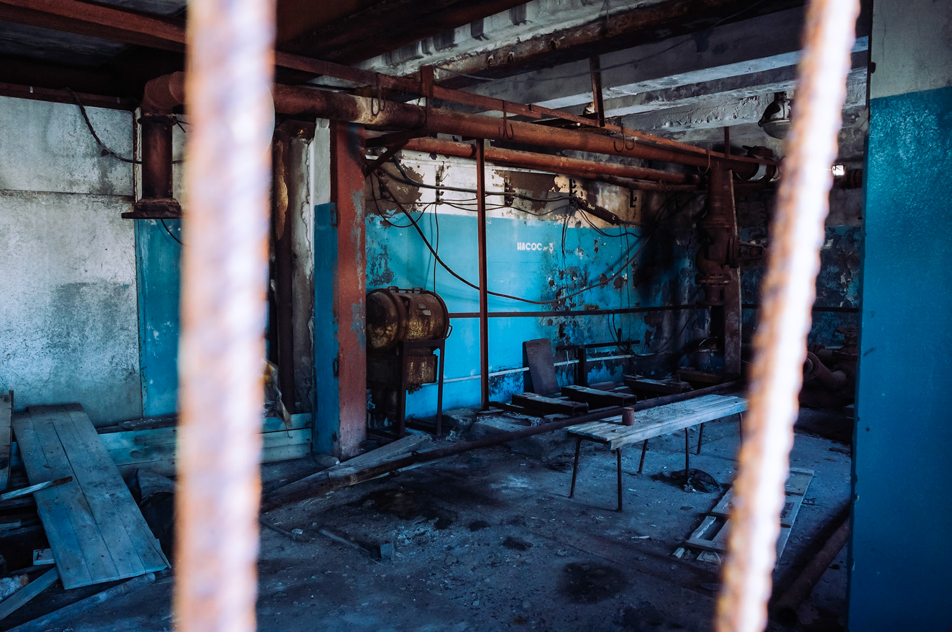 Ein Blick durch ein rostiges Gitter in das Innere eines verlassenen Gebäudes in Pyramiden, Spitzbergen. Die Wände sind blau gestrichen, während alte Rohre und Maschinen von Verfall gezeichnet sind.