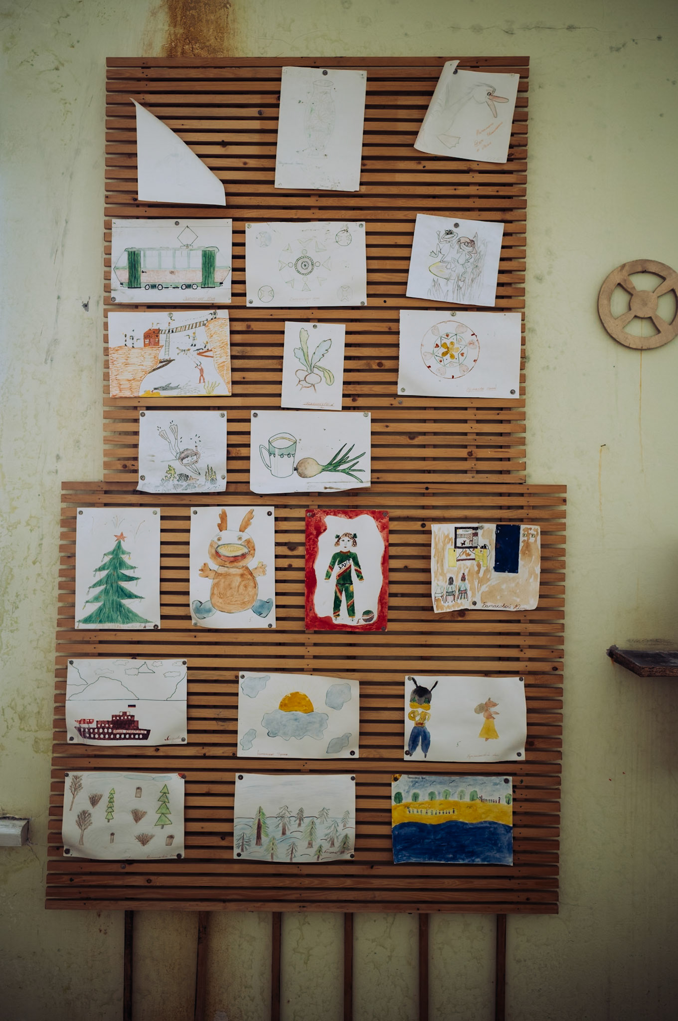 An einer Holzwand hängen zahlreiche handgemalte Kinderzeichnungen aus der Zeit, als die Schule in Pyramiden noch in Betrieb war. Die Motive reichen von Landschaften über Tiere bis hin zu fantasievollen Szenen.