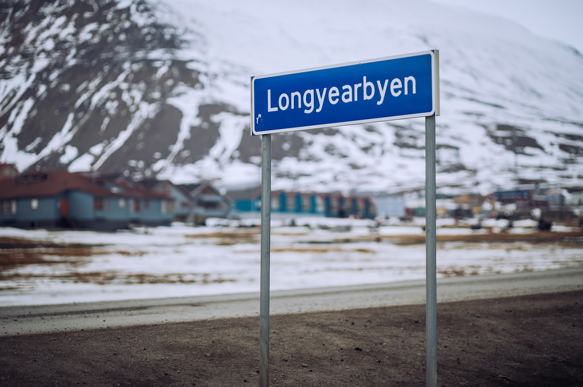 Ein Schild mit der Aufschrift 'Longyearbyen' steht am Straßenrand. Dahinter erkennt man schneebedeckte Berge und Gebäude.