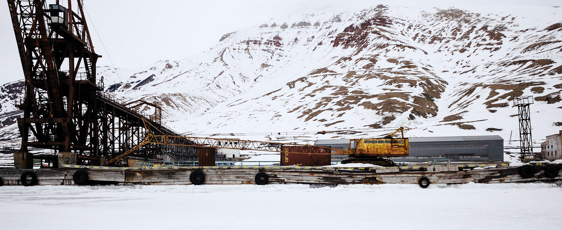 Ein verrosteter Kran und industrielle Strukturen in der verlassenen Siedlung Pyramiden. Im Hintergrund erstrecken sich die schneebedeckten Berge.