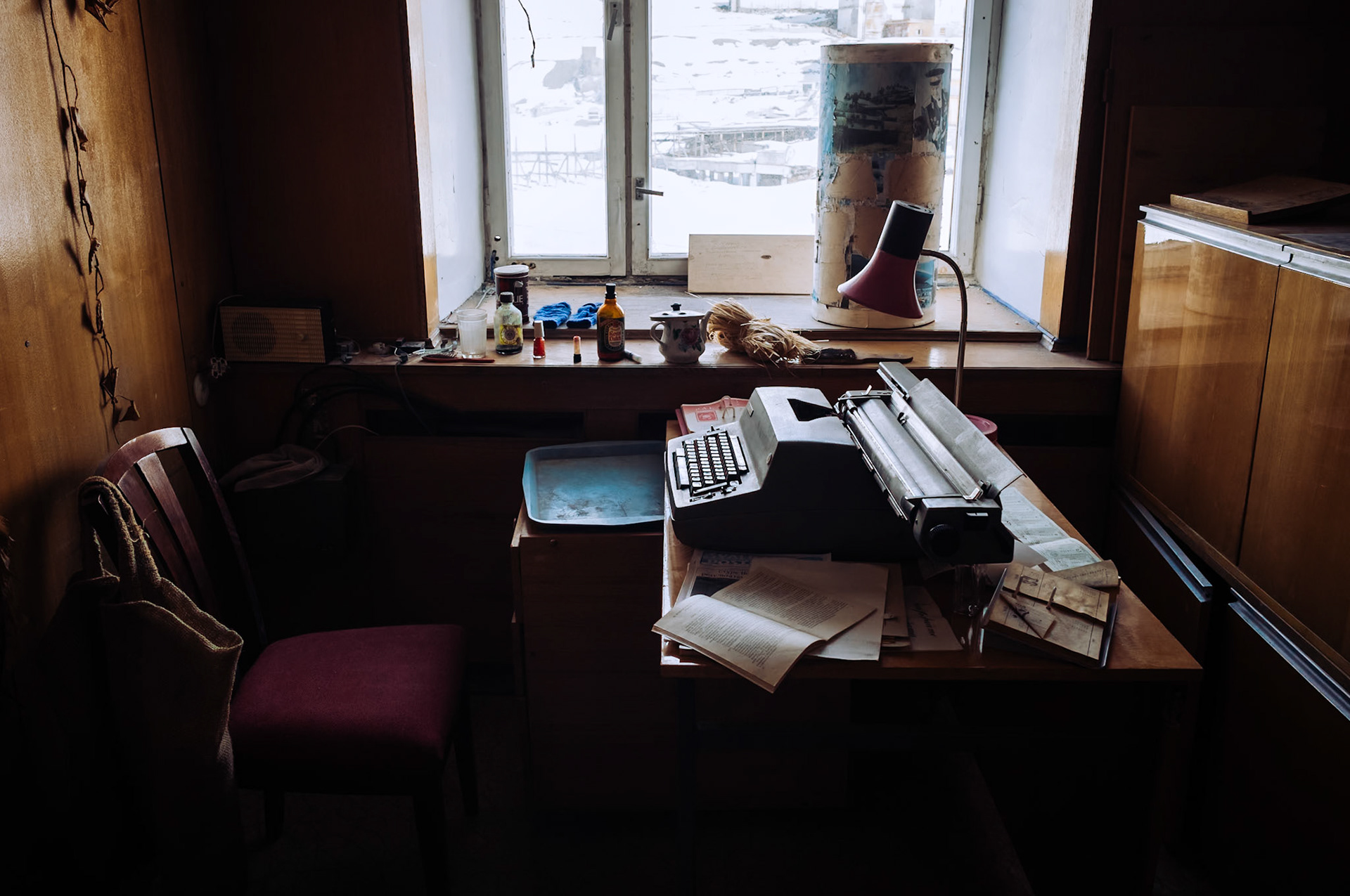 Ein Raum mit Holzvertäfelung in Pyramiden, Spitzbergen, enthält eine alte Schreibmaschine auf einem Schreibtisch neben verstreuten Dokumenten. Die vergilbten Papiere und das Fenster mit Schneeblick zeugen von der verlassenen Vergangenheit.