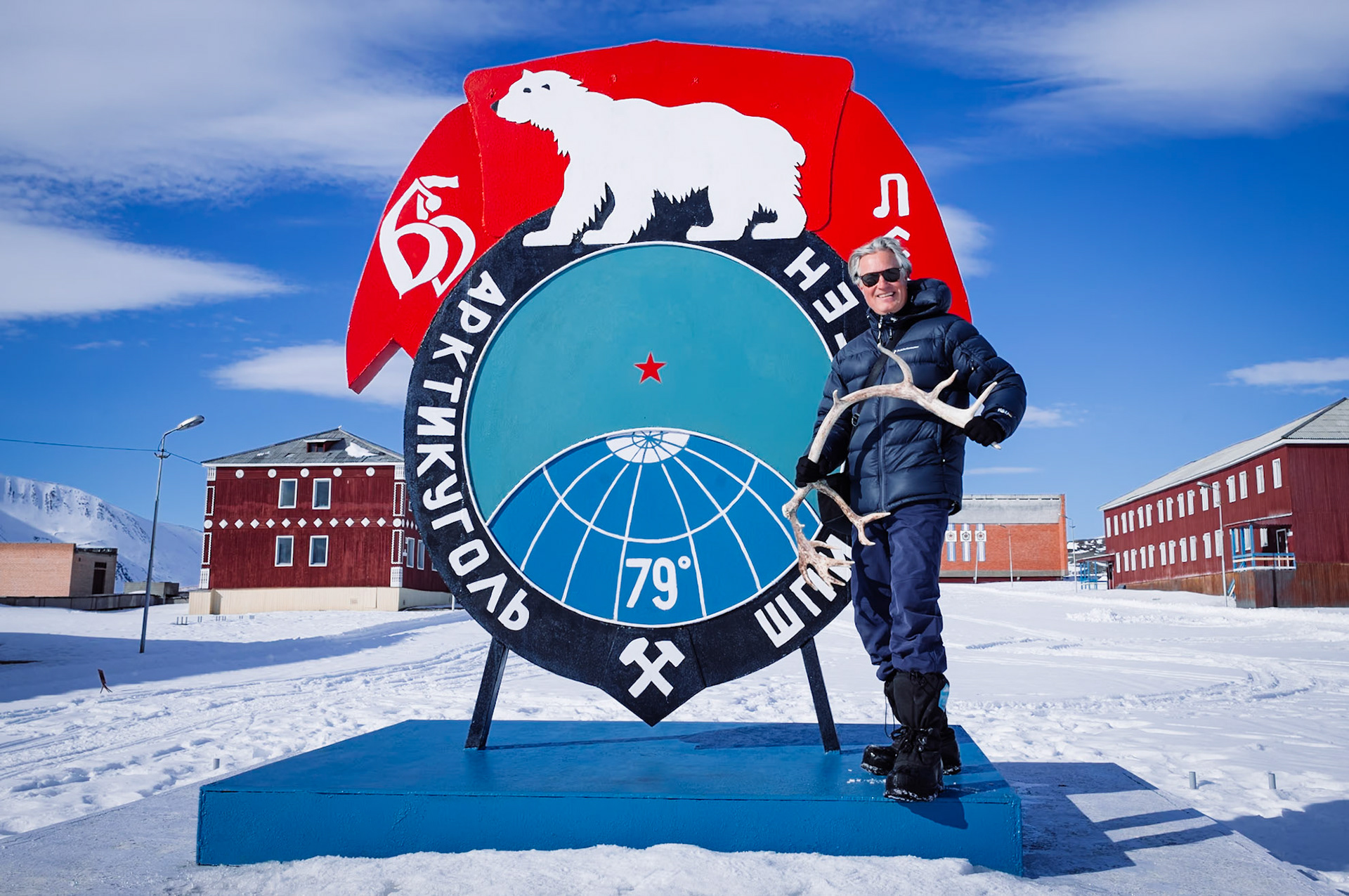 Der Autor posiert vor einem sowjetischen Denkmal mit einem Eisbärenmotiv und der Aufschrift 'Арктикуголь' in Pyramiden, Spitzbergen. Die Szene zeigt die Verbindung zur sowjetischen Bergbauvergangenheit der Siedlung.