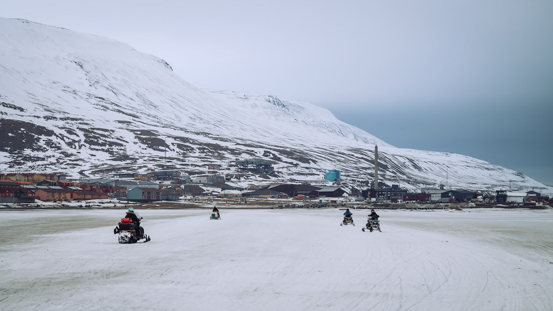 Menschen fahren mit Schneemobilen durch die schneebedeckte Landschaft von Longyearbyen. Im Hintergrund sind bunte Häuser und Berge zu sehen.