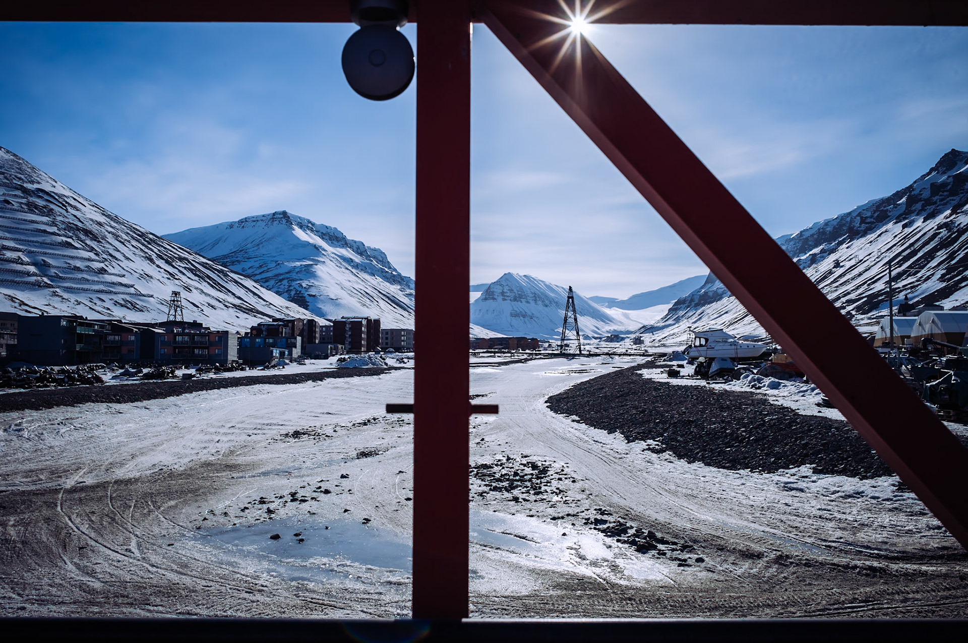 Eine Ansicht von Longyearbyen, eingerahmt durch rote Stahlträger, die zum Teil eines Gebäudes oder einer Brücke gehören. Im Hintergrund sind schneebedeckte Berge und Gebäude der Siedlung zu sehen.