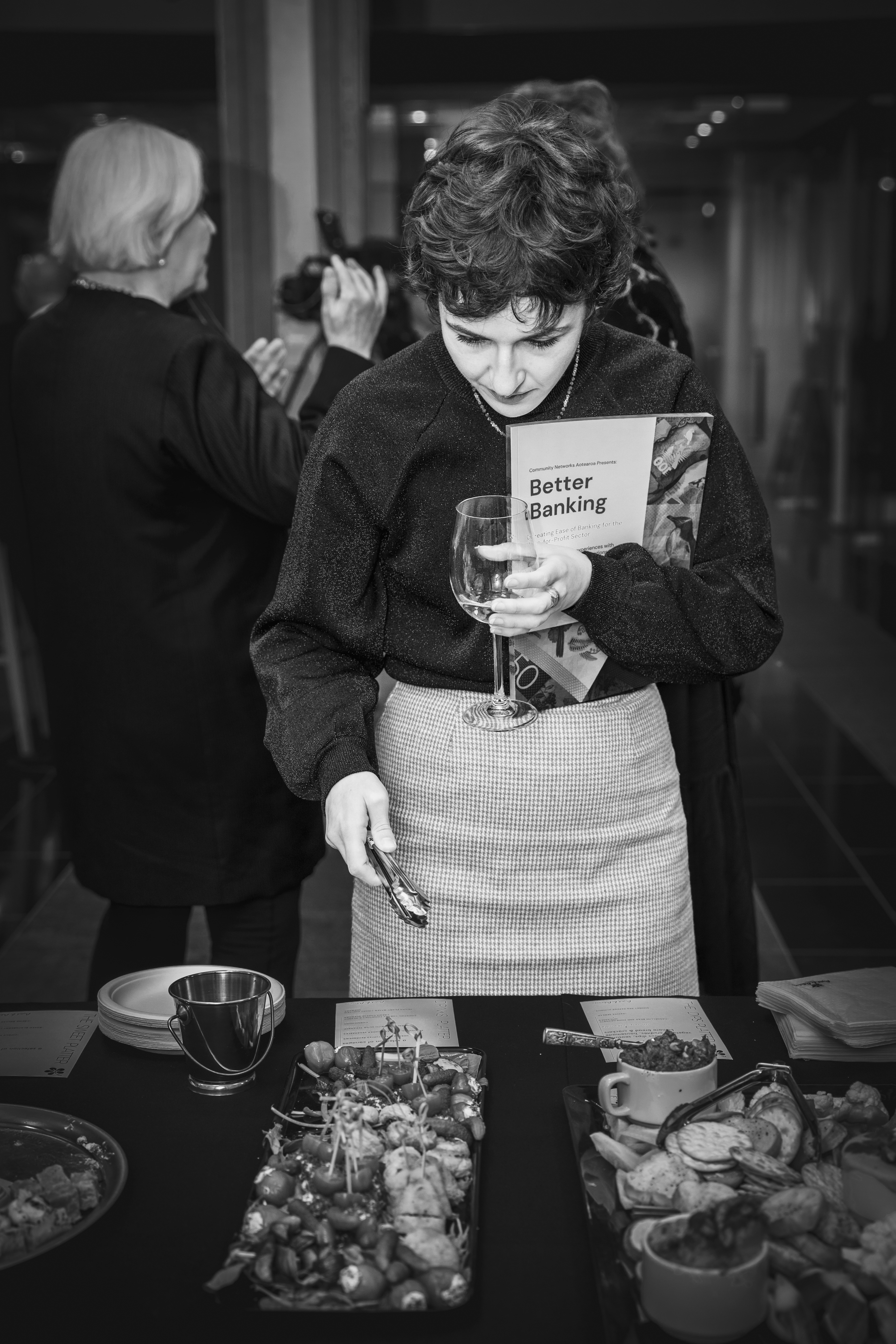 An attendee holds a glass of wine and selects food while holding a Better Banking booklet close to themselves.