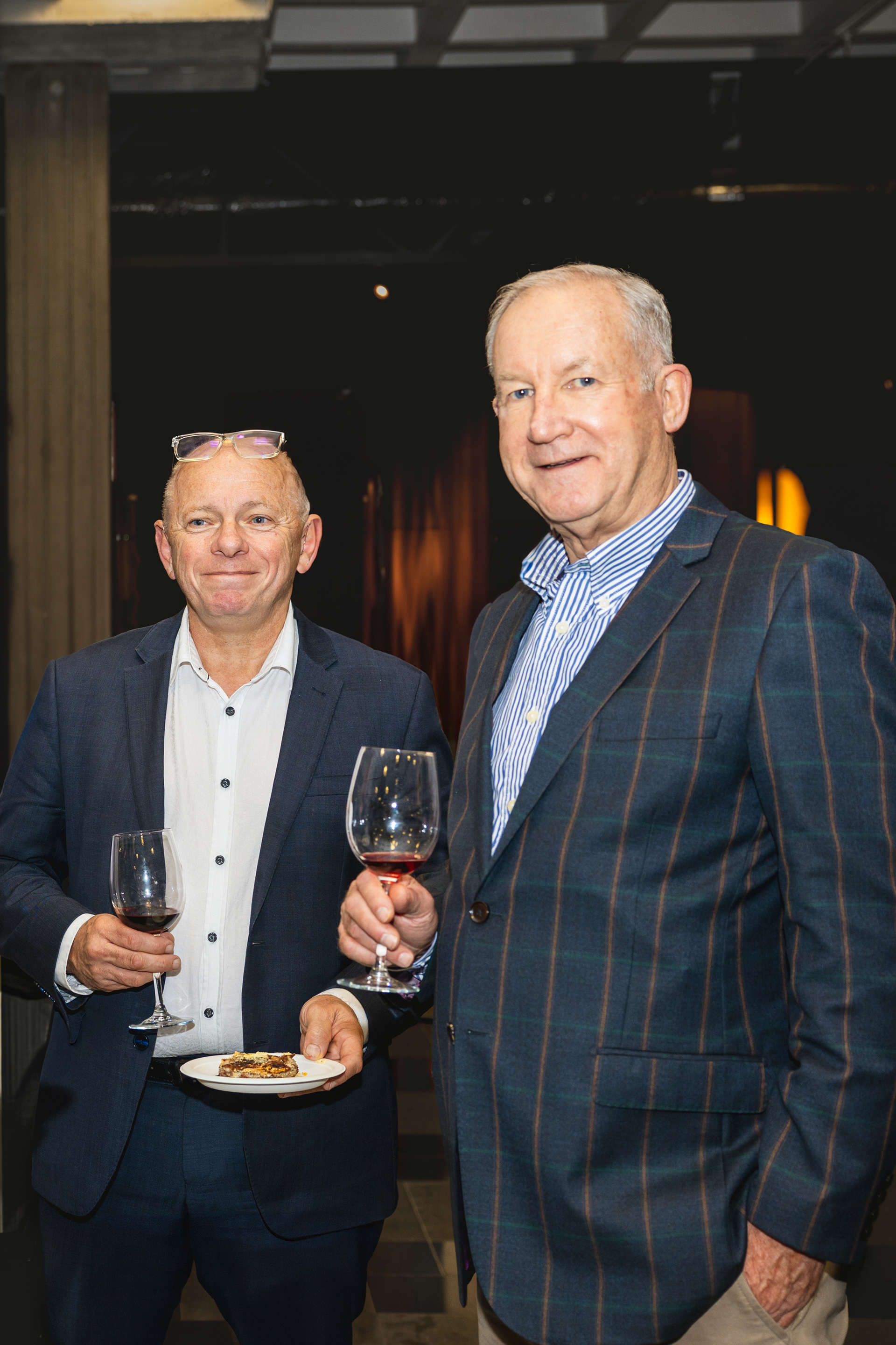 Two men in suits smile and pose for a photo while holding glasses of red wine.