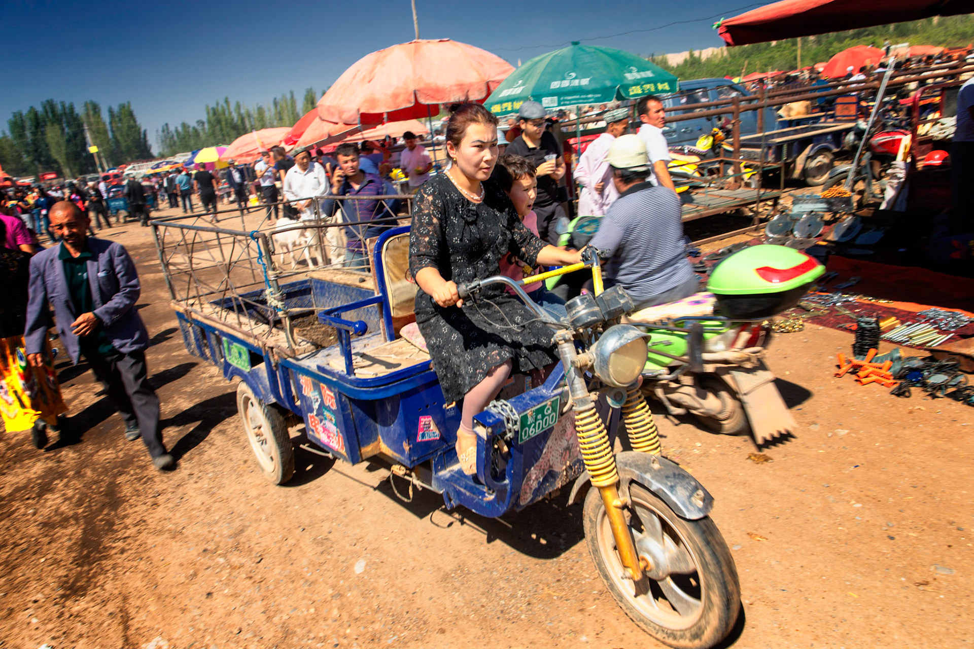 Kashgar,  China - September 9th, 2018 : Woman driving a three-wheeler at the cattle market, people in and parasols int the background.